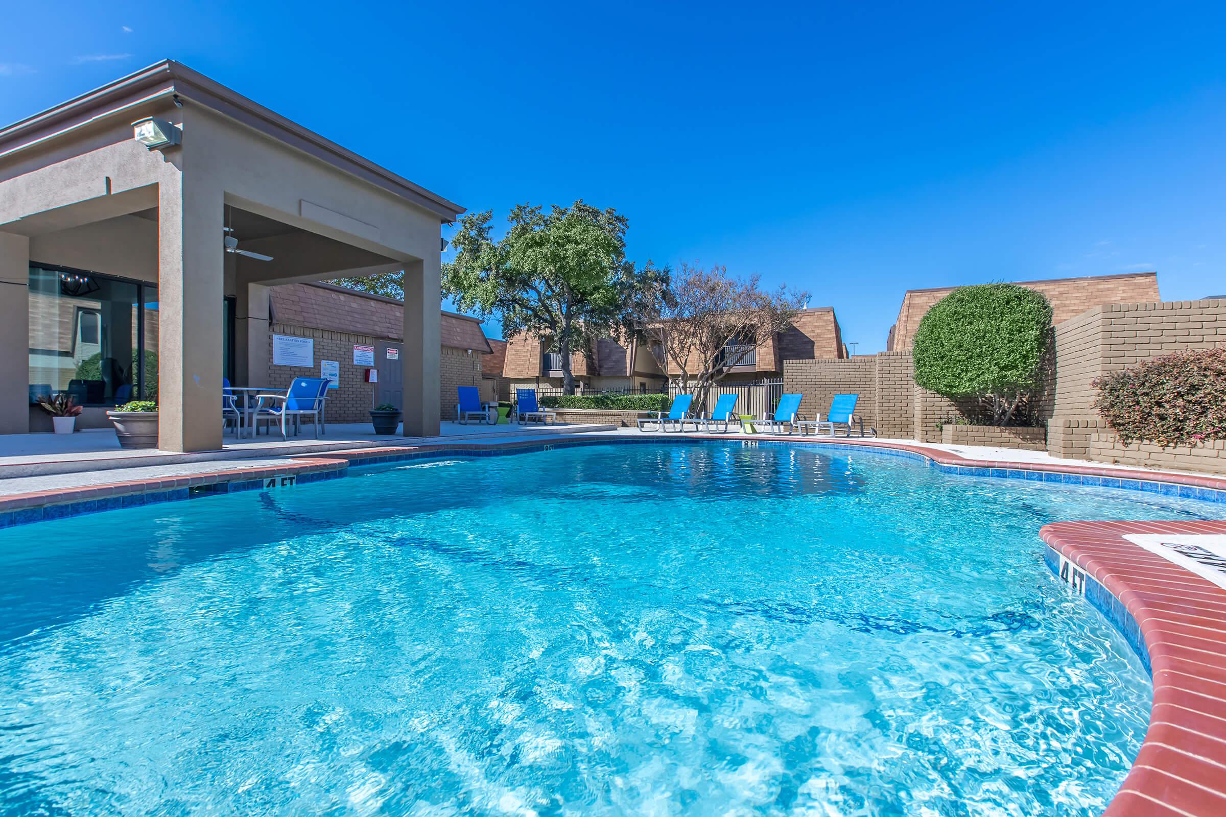 A clear swimming pool with blue water surrounded by lounge chairs and a shaded area. In the background, there are landscaped trees and brick walls. The sky is bright blue, creating a sunny and inviting atmosphere.