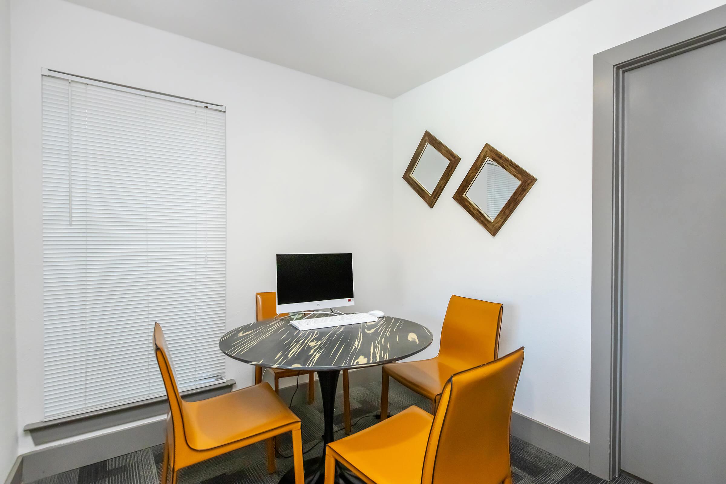 A small, modern workspace featuring a round black and white table with a computer on it. Four orange chairs surround the table. The room has white walls, a window with blinds, and two decorative mirrors hanging on the wall. A gray door is visible in the background.