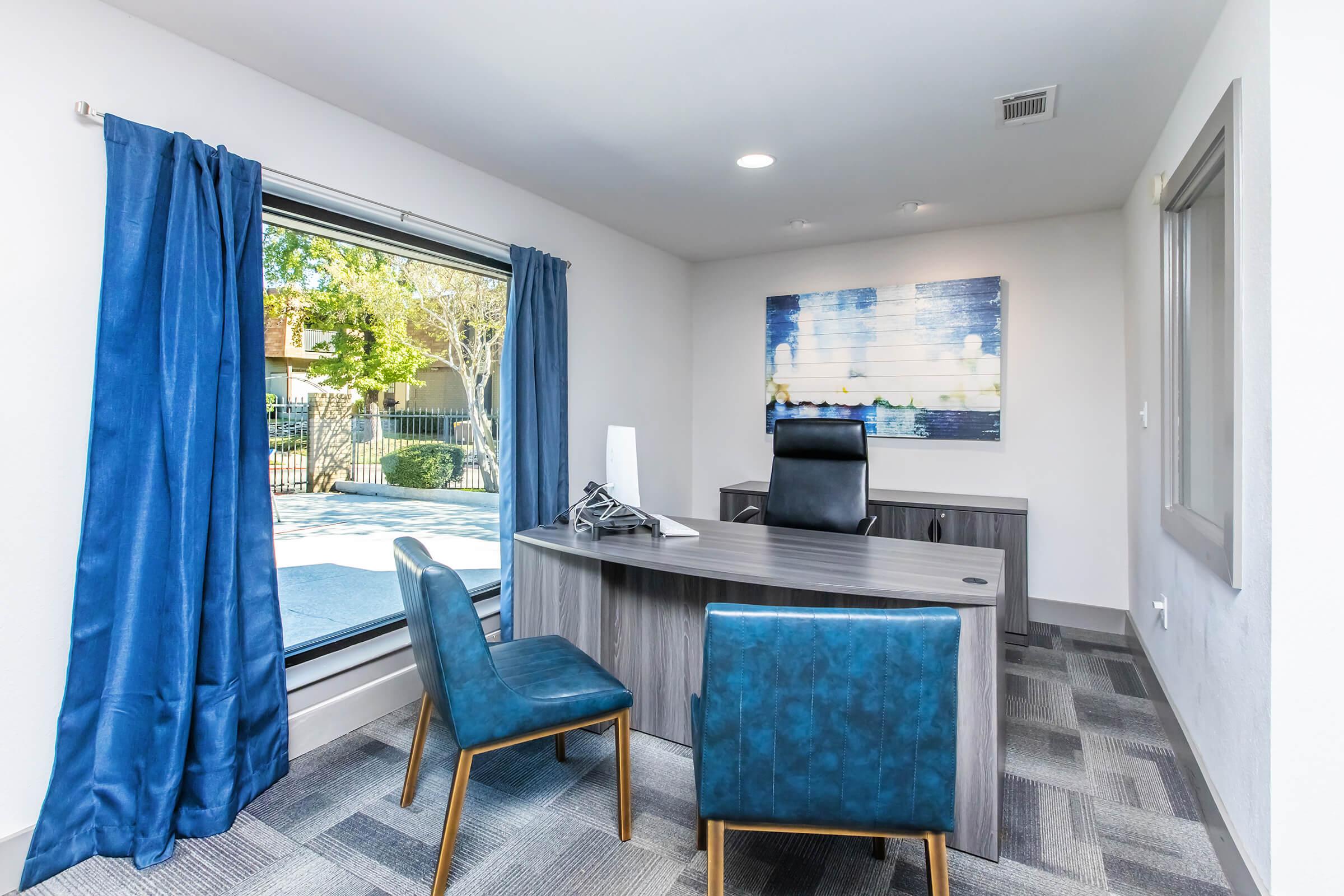 A modern office space featuring a sleek wooden desk with a black ergonomic chair. Two blue upholstered chairs are positioned in front of the desk. The room has large windows with blue curtains, providing natural light and a view of greenery outside. The flooring is gray carpet with a contemporary pattern.