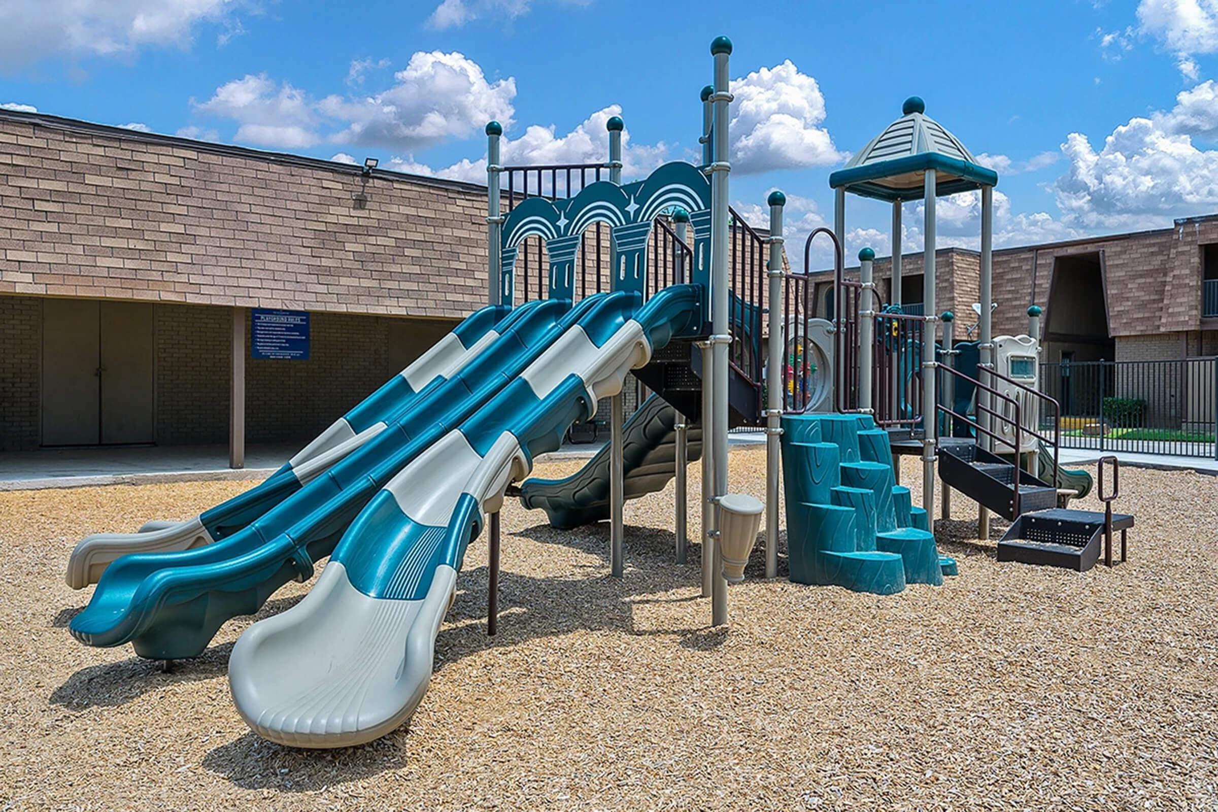 A colorful playground featuring multiple slides of varying heights and lengths, surrounded by a layer of light-colored gravel. The structure includes steps leading up to the slides, a small covered area, and safety railings. The sky is bright with fluffy clouds in the background.