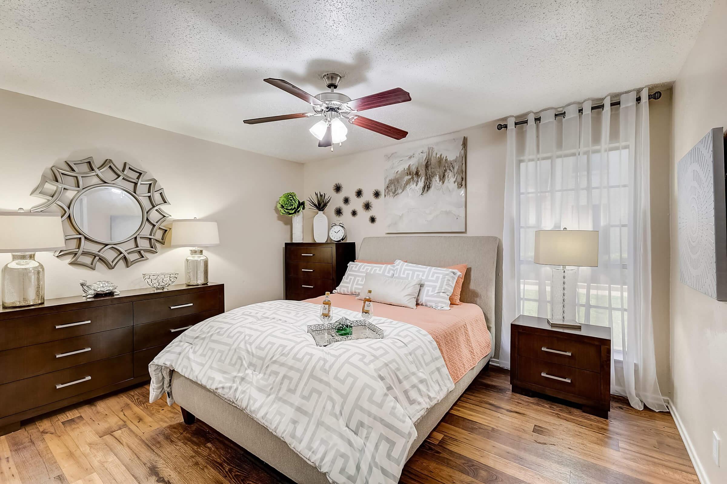 A cozy bedroom featuring a queen-sized bed with a light orange and white patterned bedspread. There are two dark wooden nightstands with lamps on either side, and a decorative mirror above a dresser. A large window with sheer curtains allows natural light to enter, complemented by stylish wall art and a fan overhead.