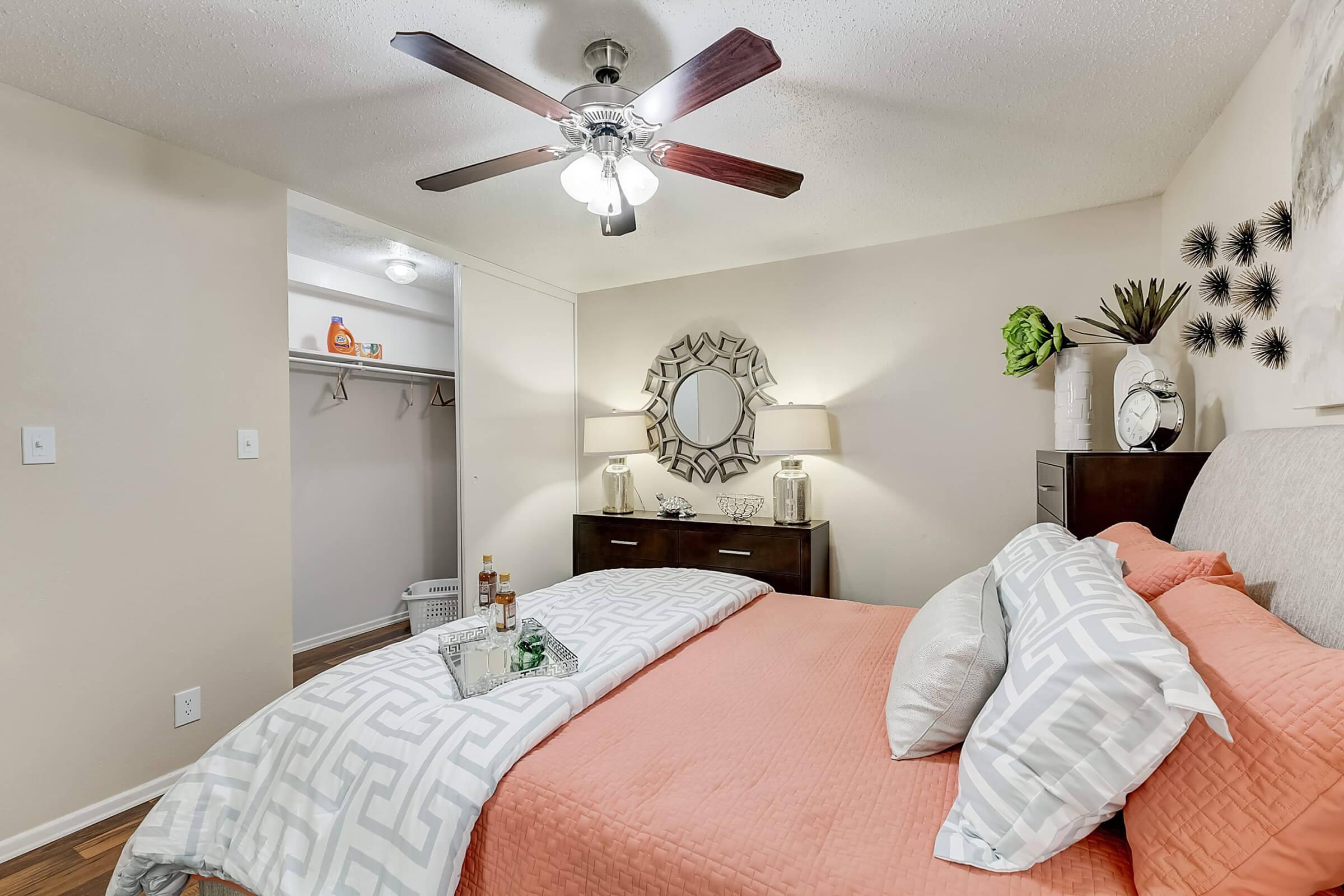 A cozy bedroom featuring a queen-sized bed with a coral quilt and decorative pillows. A ceiling fan spins overhead, and a large mirror adorns the wall. A dark wooden dresser and a closet are visible, with some decorative plants and bottles on the nightstand, adding a warm ambiance to the space.