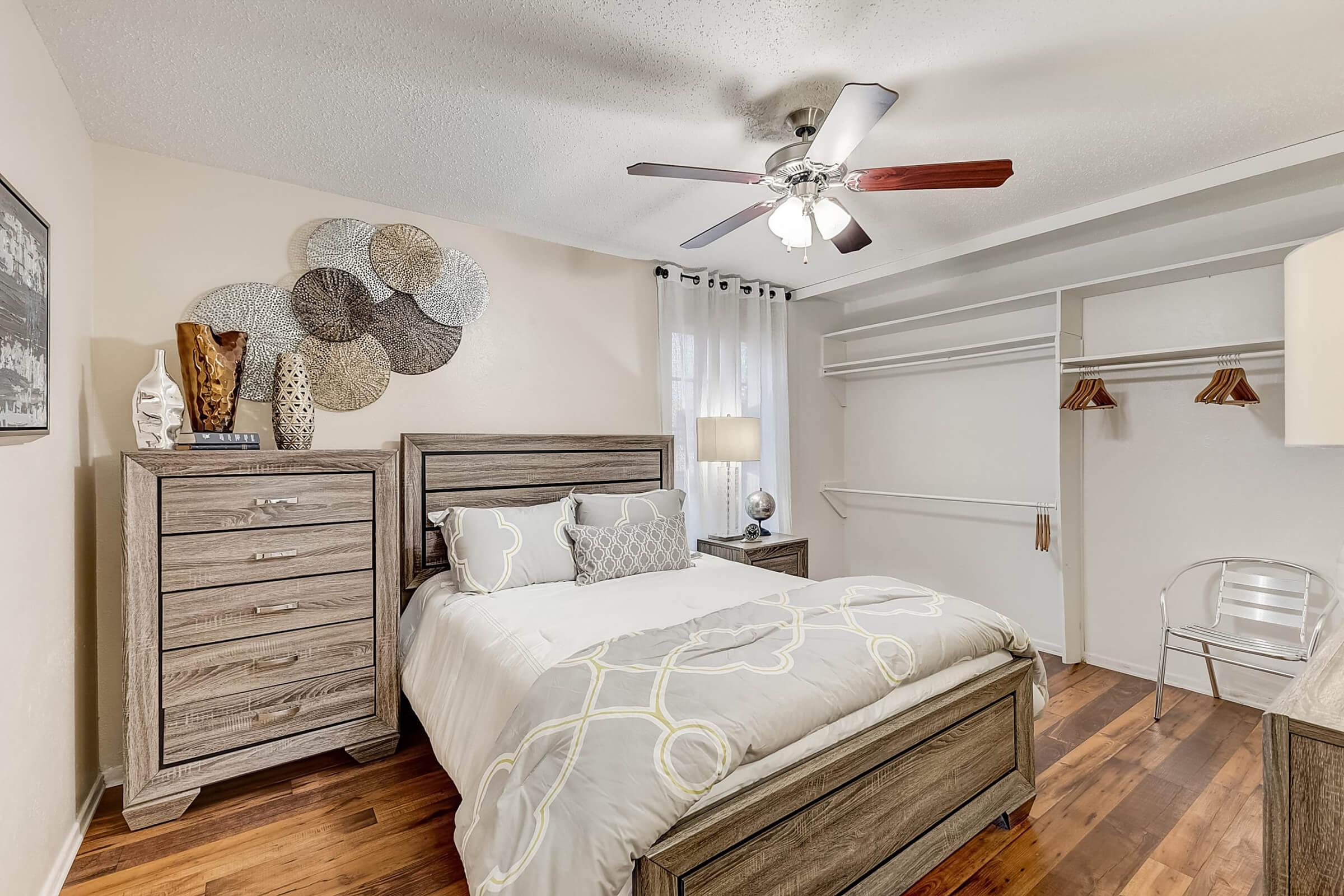 A cozy bedroom featuring a queen-sized bed with decorative pillows, a wooden dresser, and a ceiling fan. The room has stylish woven wall decor, a floor lamp, and a spacious closet with hangers. Soft natural light filters through a window with sheer curtains, enhancing the inviting atmosphere.