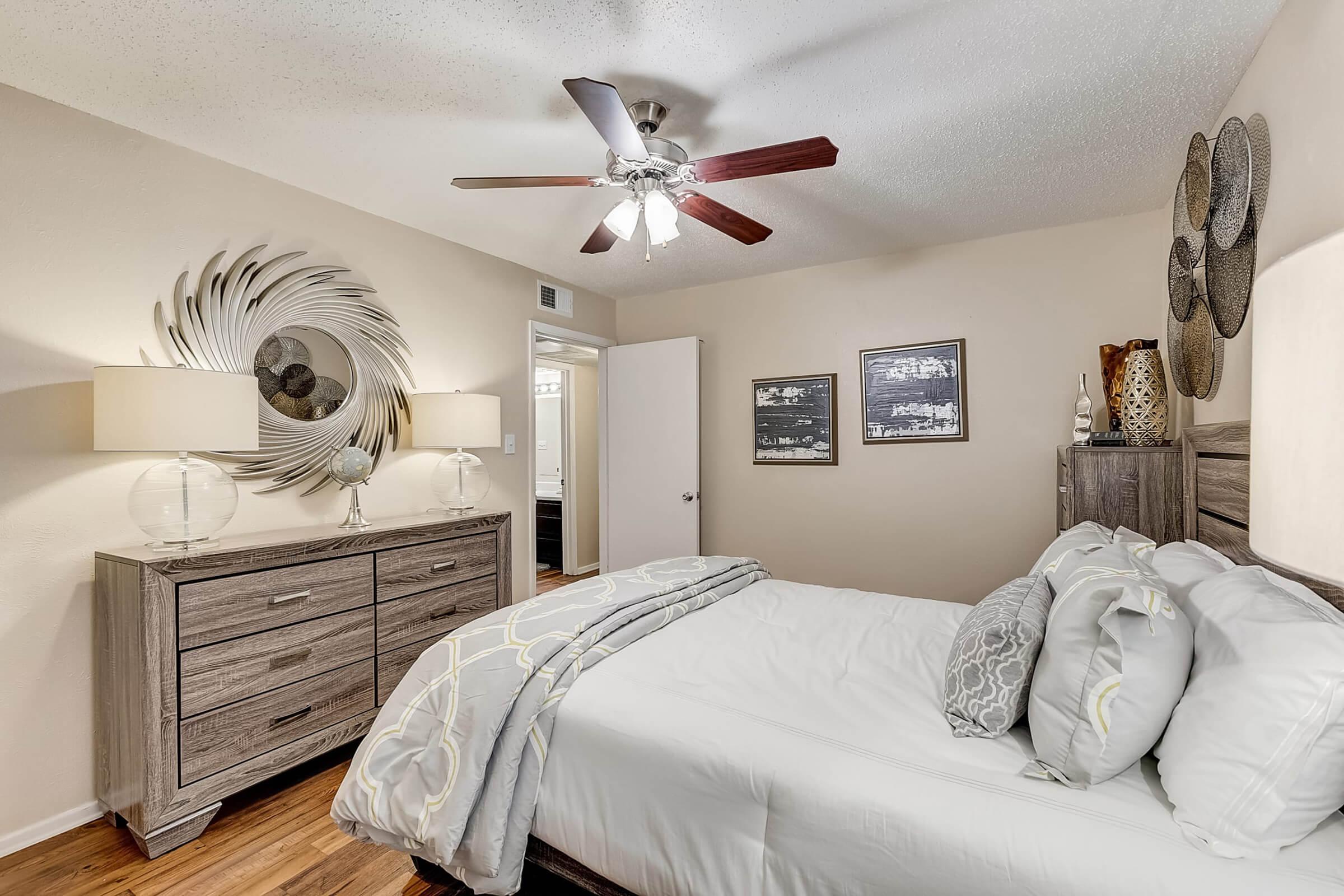 A cozy bedroom featuring a queen-sized bed with decorative pillows, a modern nightstand, and a wooden dresser. The room includes a ceiling fan, wall art, and a circular mirror. Soft lighting creates a warm atmosphere, complemented by light-colored walls and wooden flooring.