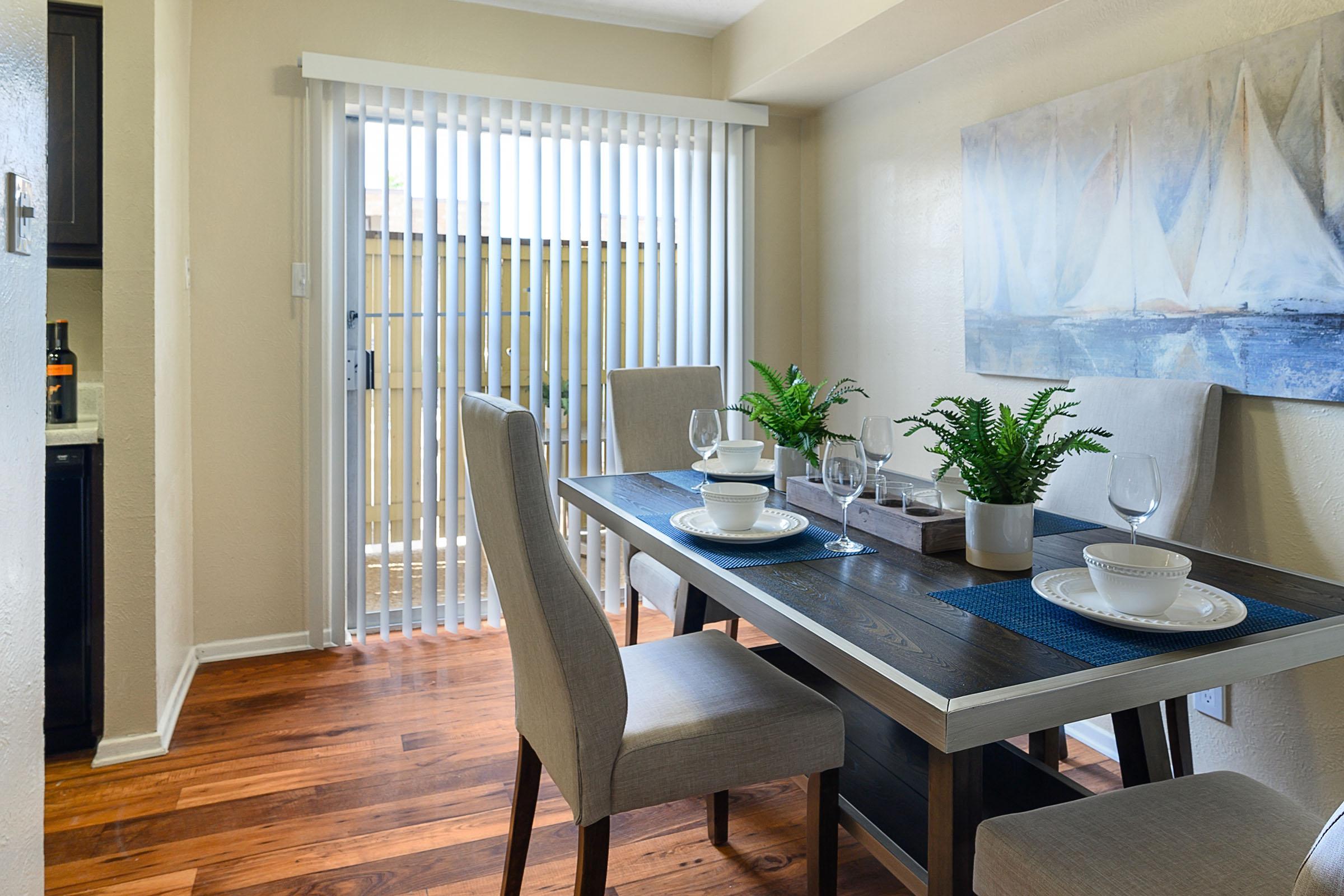 A bright dining area featuring a wooden table set with dishes and glassware. Four elegant beige chairs surround the table, and a green plant decorates the center. Vertical blinds are partially drawn at a sliding glass door, leading to an outdoor space. A serene sailboat painting adorns the wall.