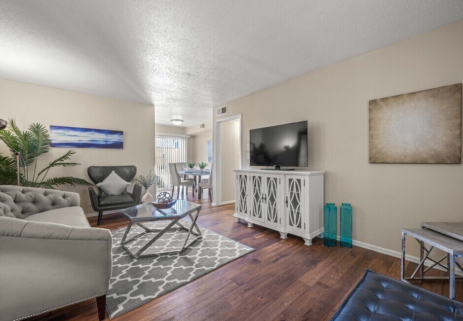 A cozy living room featuring a light beige color scheme, a tufted gray couch, and a glass coffee table. There's a white cabinet with decorative patterns, a black leather ottoman, and a flat-screen TV. A dining area with a wooden table is visible in the background, and sunlight streams in through window blinds.