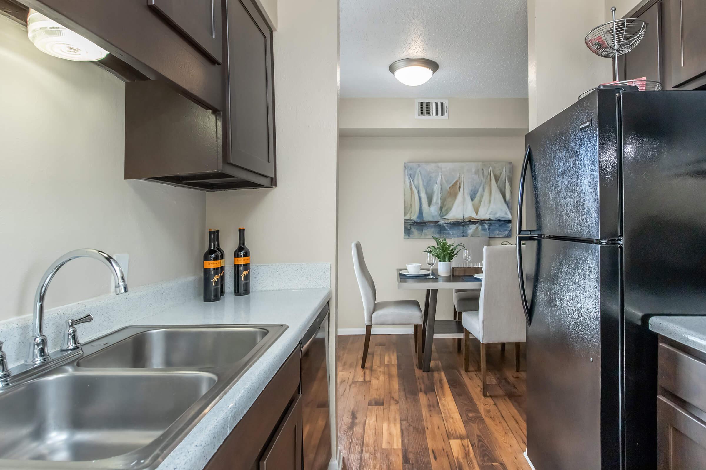 A modern kitchen featuring dark wood cabinetry, a double sink, and a black refrigerator. Two bottles of wine are displayed on the counter. In the background, a dining area with a small table and beige chairs is visible, along with a painting of sailboats on the wall. The flooring is wood-like laminate.