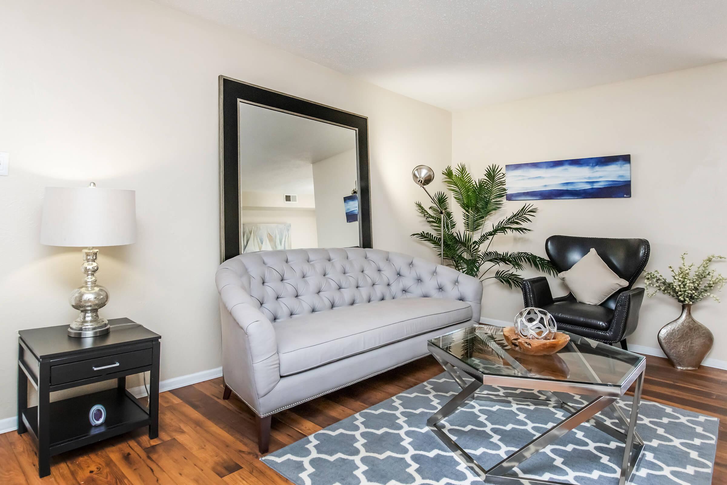 A cozy living room featuring a light gray tufted sofa, a sleek black armchair, and a mirrored wall. A modern glass coffee table sits on a patterned rug, accompanied by a decorative bowl. A stylish lamp and potted plant add warmth to the space, with a piece of abstract art in the background.