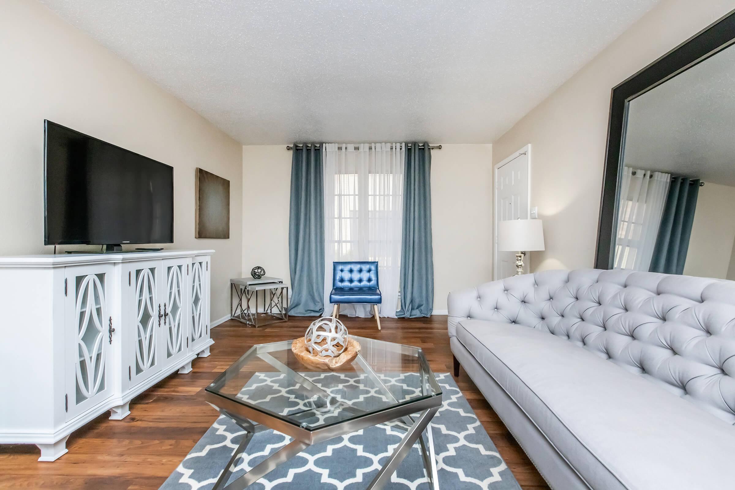 A stylish living room featuring a light gray tufted sofa, a glass coffee table with a decorative centerpiece, and a large mirror. A modern TV stands on a white entertainment unit, with dark blue curtains and a blue accent chair in the background, enhancing the contemporary decor.