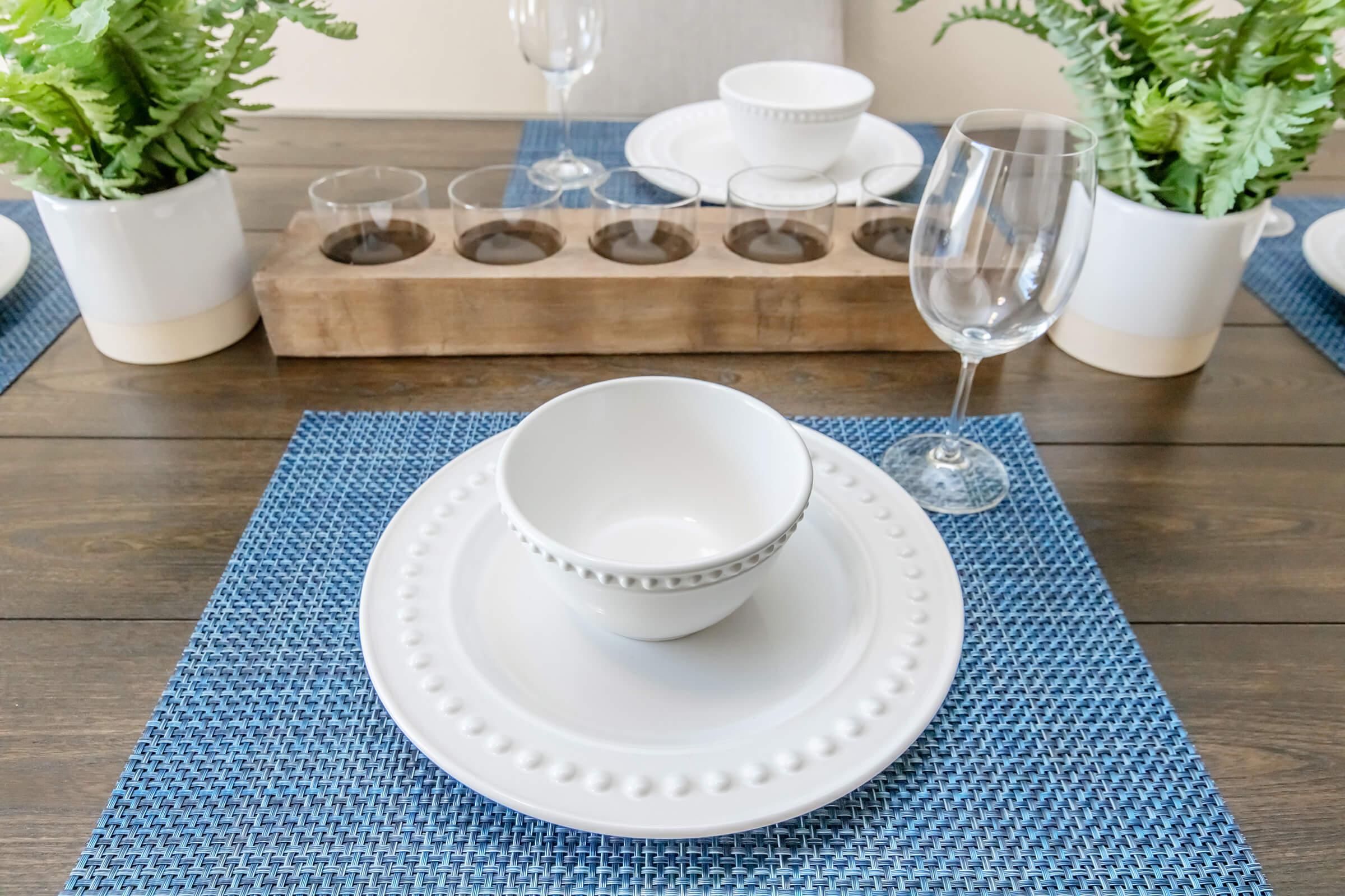 A beautifully arranged dining table featuring white dinnerware with decorative beading, a central bowl, drinking glasses, and two wine glasses. The table is set with a blue woven placemat and includes potted ferns as centerpieces, all resting on a wooden table.