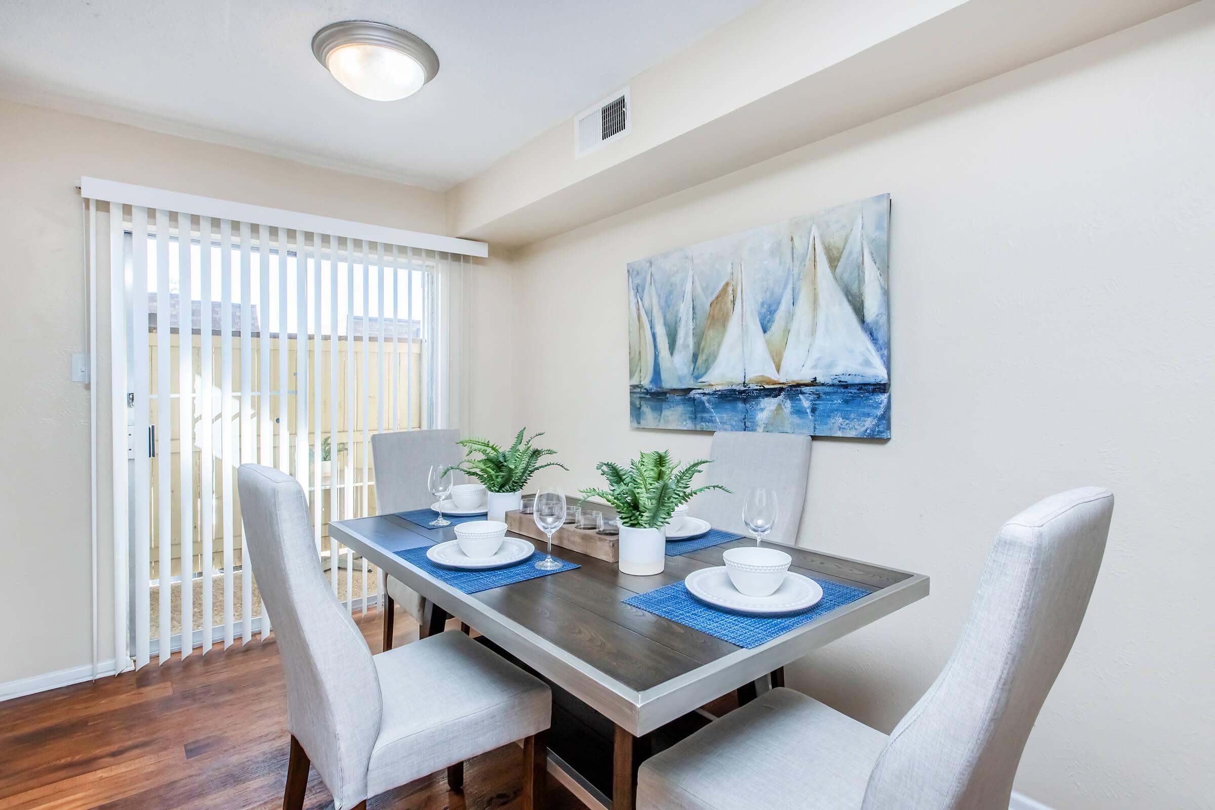 A dining area featuring a wooden table set for four with white plates, glasses, and blue placemats. There are two light-colored upholstered chairs on either side. A painting of sailboats hangs on the wall, and large sliding doors with vertical blinds provide natural light. A small potted plant is in the center of the table.