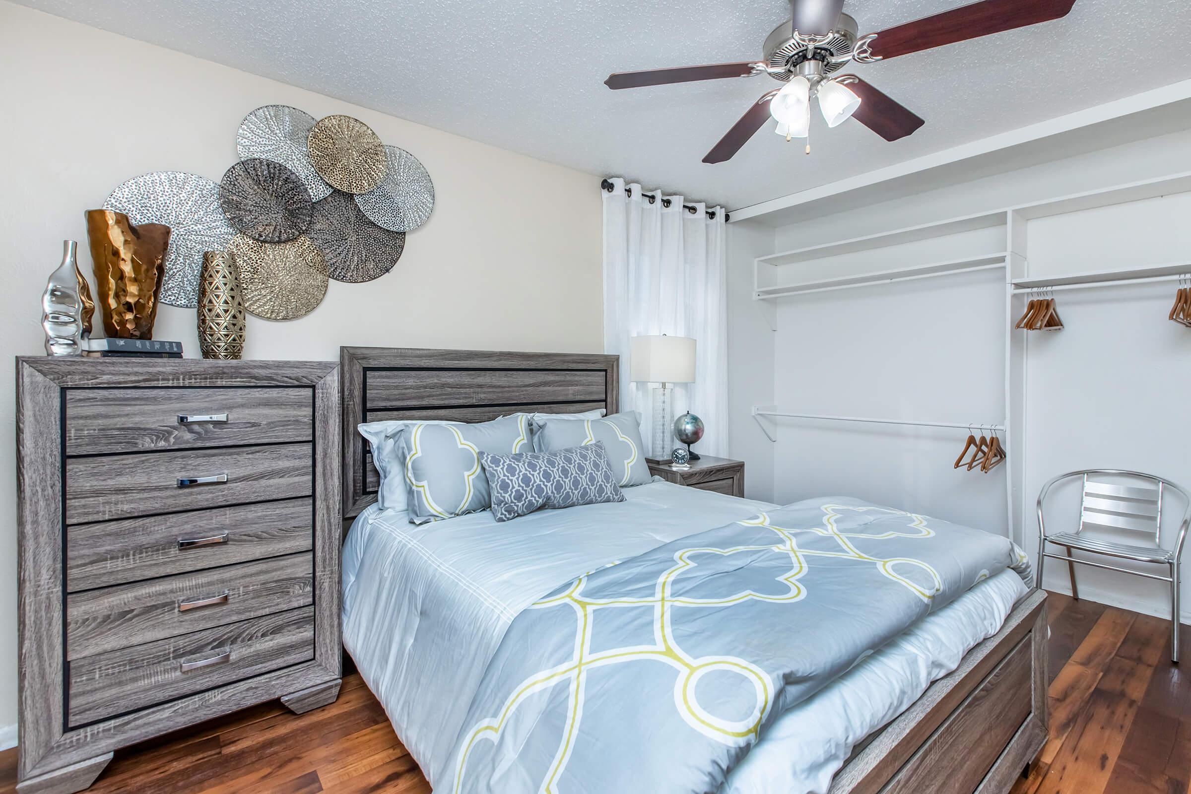 A cozy bedroom featuring a wooden bed frame with a light blue and white patterned duvet, a nightstand with a lamp, and a tall dresser. The wall is decorated with circular metal wall art, and the room has a ceiling fan. A closet with hanging space is visible, showcasing a clean and elegant design.