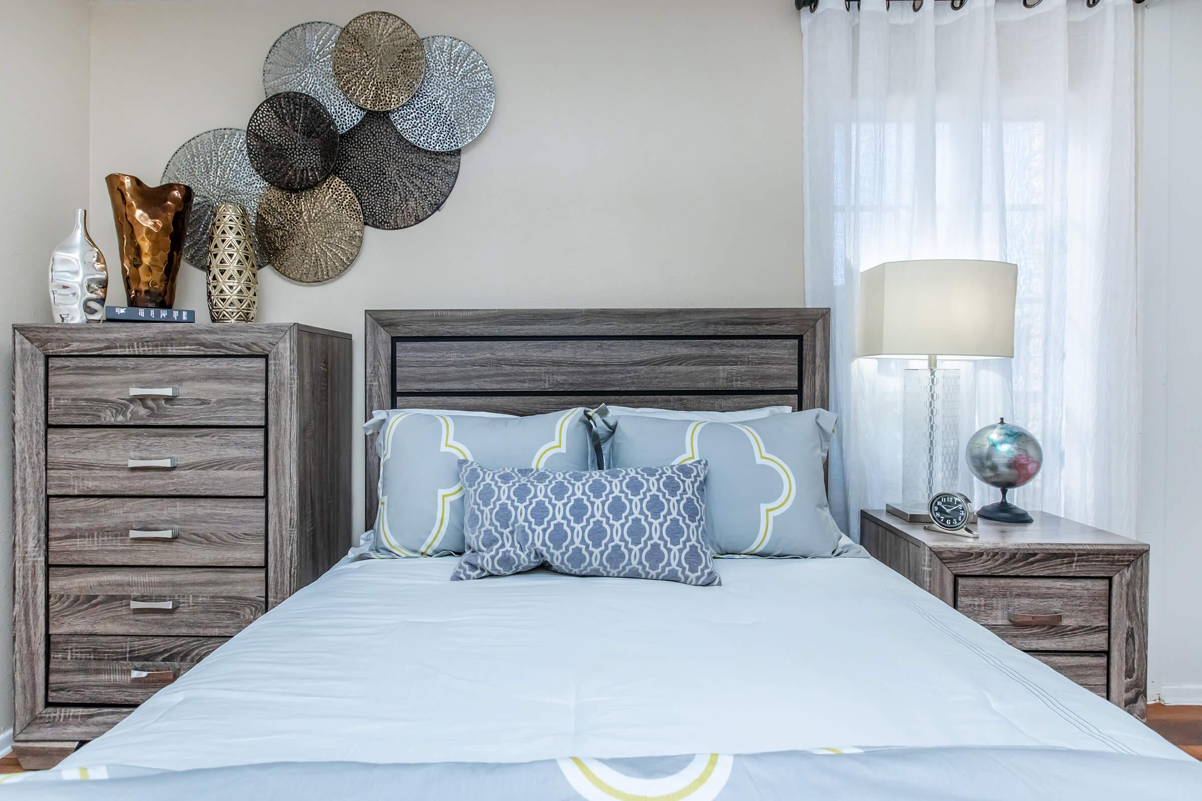 A neatly made bed with light blue bedding and decorative pillows, flanked by wooden nightstands and a tall dresser. The wall features a decorative arrangement of metallic wall art, and there’s a lamp on the nightstand. Natural light filters through sheer curtains, creating a cozy atmosphere.