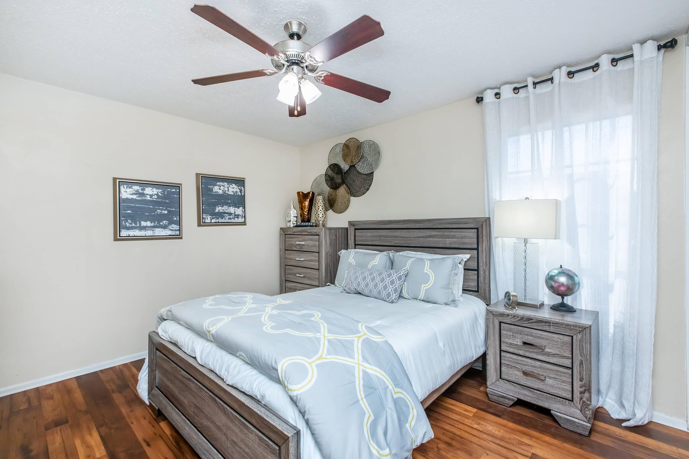 A cozy bedroom featuring a queen-sized bed with a light blue comforter and decorative pillows. A wooden nightstand with a lamp sits beside the bed, and a dresser is against the wall. The room has light-colored walls, hardwood flooring, and a ceiling fan. Two framed pictures are hung on the wall.