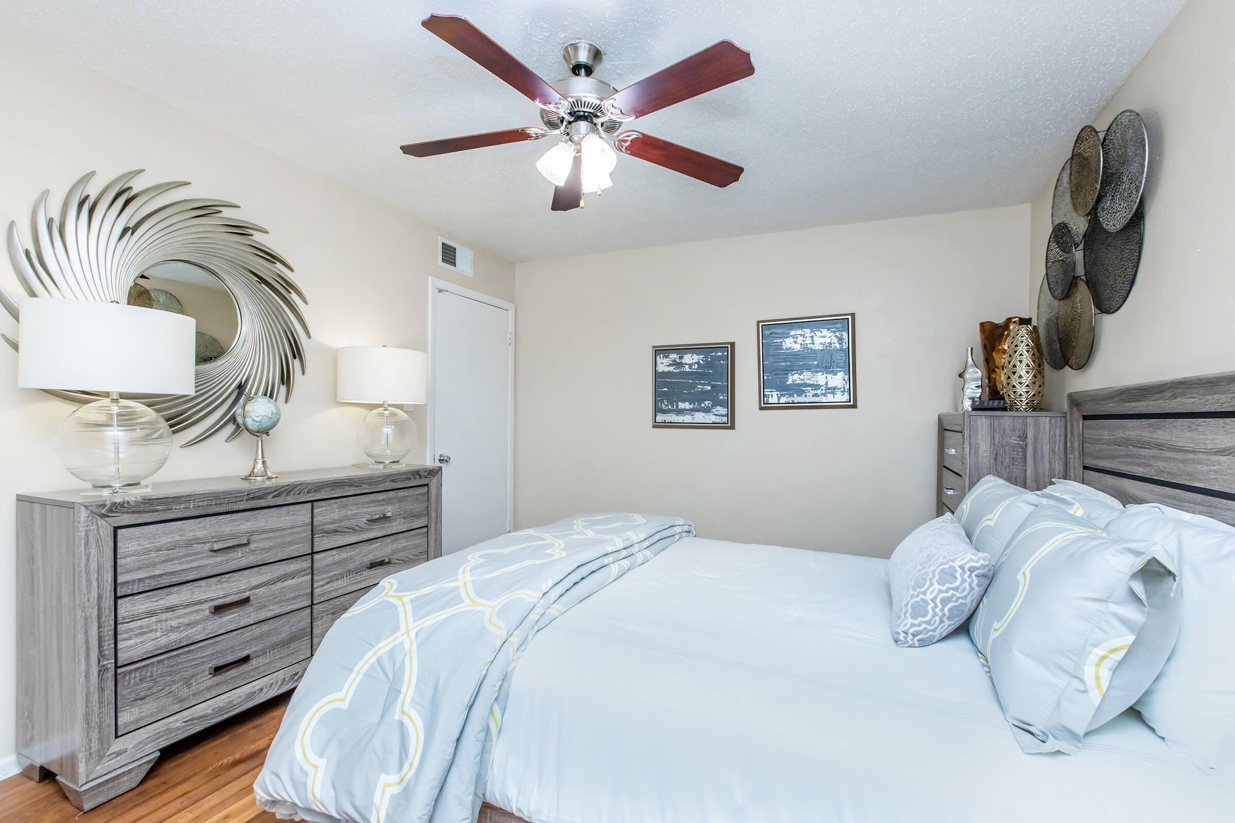 A neatly arranged bedroom featuring a queen-sized bed with light gray bedding and decorative pillows. A wooden dresser and nightstand accompany the bed, with a large mirror and two framed wall art pieces. A ceiling fan hangs above, and warm wooden flooring adds to the inviting atmosphere.