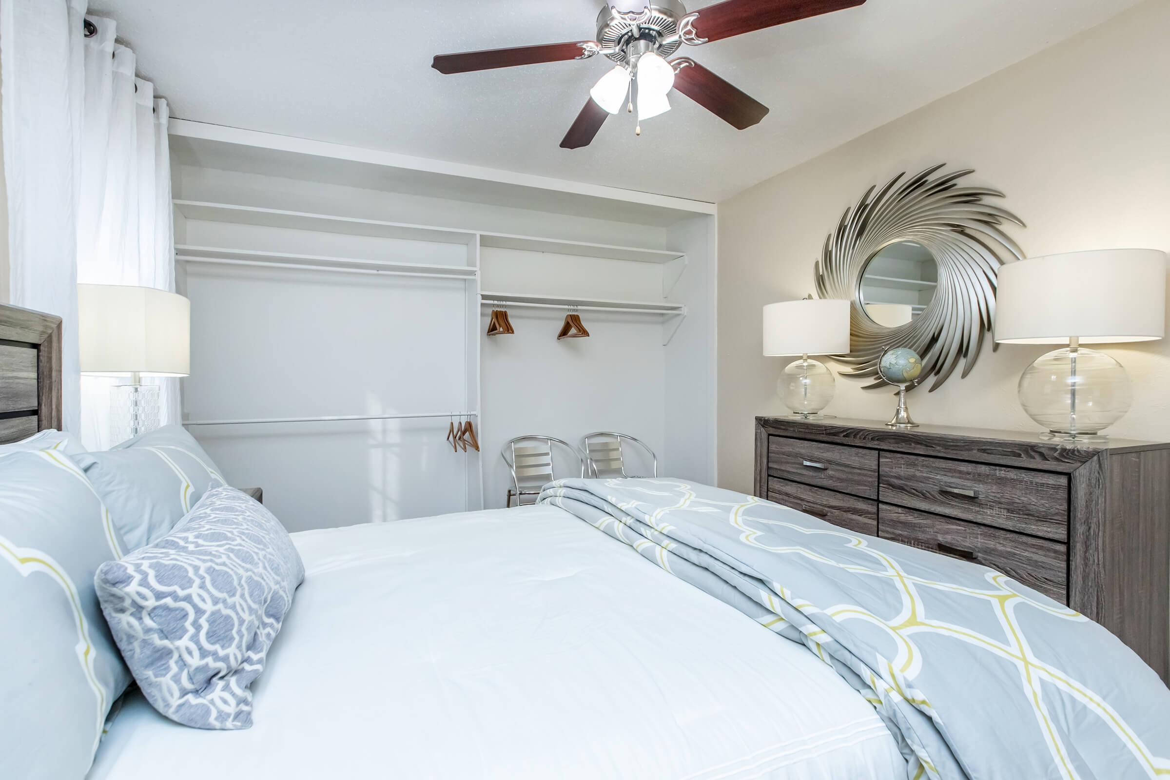 A cozy bedroom featuring a queen-sized bed with light blue bedding, two bedside lamps, and a stylish mirror on the wall. A dresser sits against one wall, while a closet with hanging space is visible. Soft, neutral colors create a calm atmosphere, enhanced by a ceiling fan.