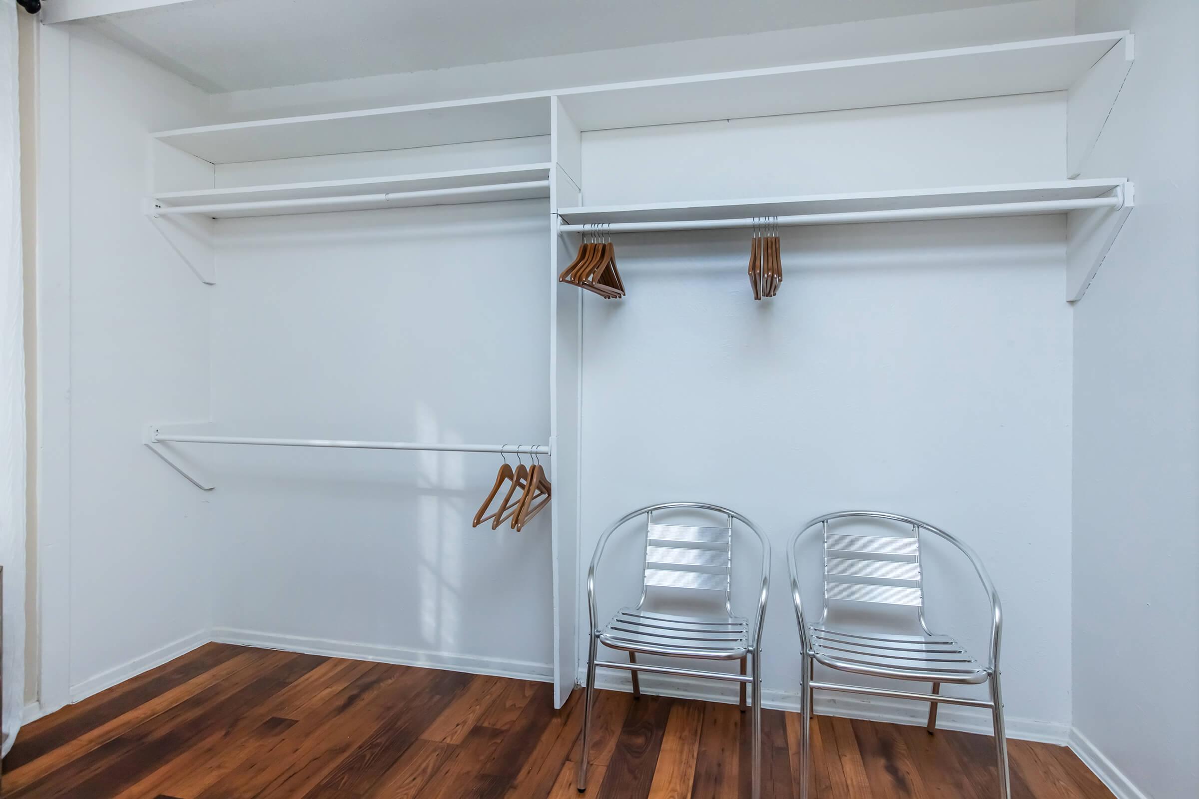 A minimalist closet featuring white shelves and hanging rods, with a few wooden hangers. Two metal chairs are positioned on a wooden floor, creating a spacious and tidy appearance. The walls are painted white, enhancing the bright and airy feel of the space.