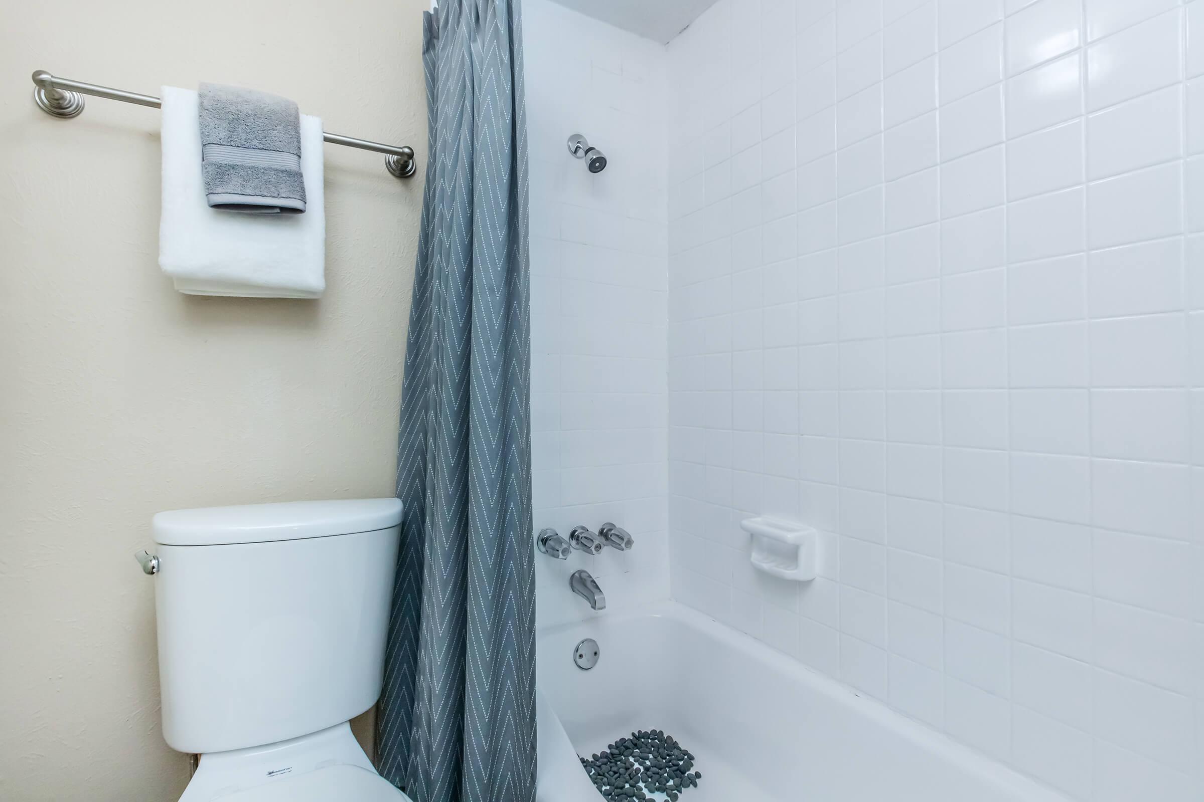 A clean, minimal bathroom featuring a white tiled shower area with a gray curtain, a toilet, and a towel rack holding a folded gray towel and a white towel.