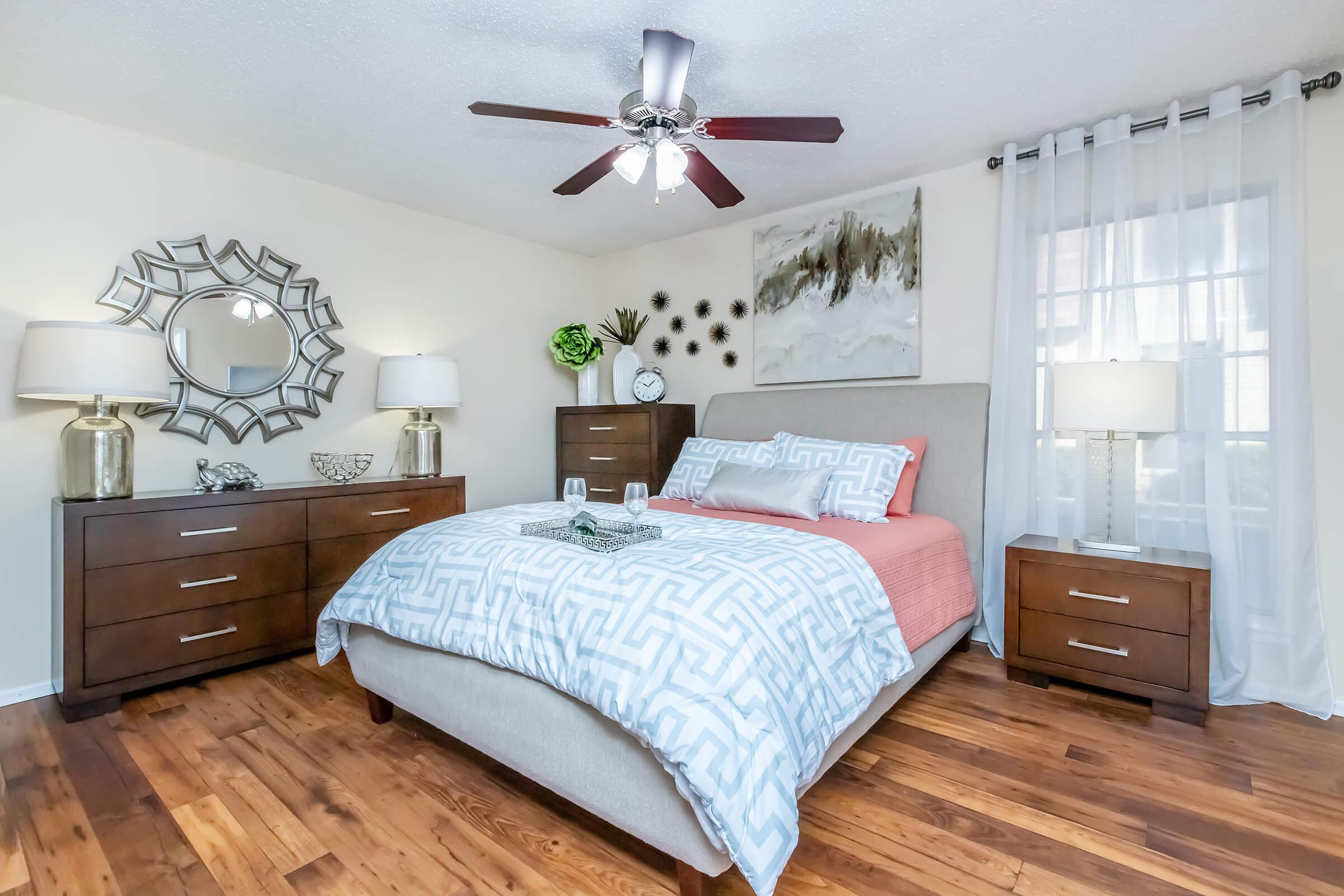 A cozy bedroom featuring a queen-sized bed with a gray and white patterned comforter, accented by coral pillows. Two wooden nightstands with modern lamps flank the bed. A decorative mirror and wall art enhance the room, while a ceiling fan adds a stylish touch. Natural light streams in through a window with sheer curtains.