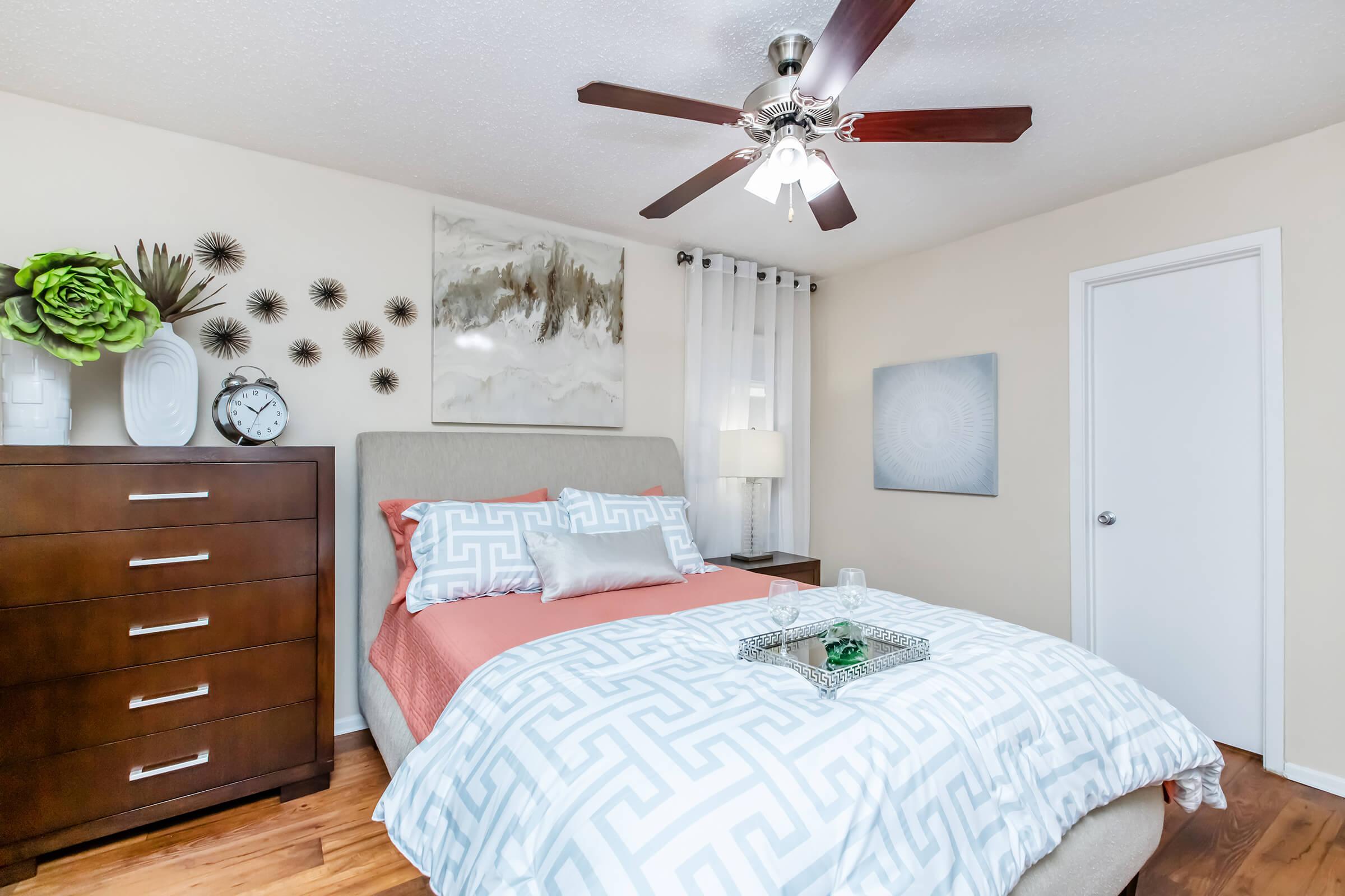 Cozy bedroom featuring a queen-sized bed with a patterned duvet in soft colors, a wooden dresser, decorative wall art, and a ceiling fan. Natural light filters through curtains, and plants add a touch of greenery, creating a warm and inviting atmosphere.