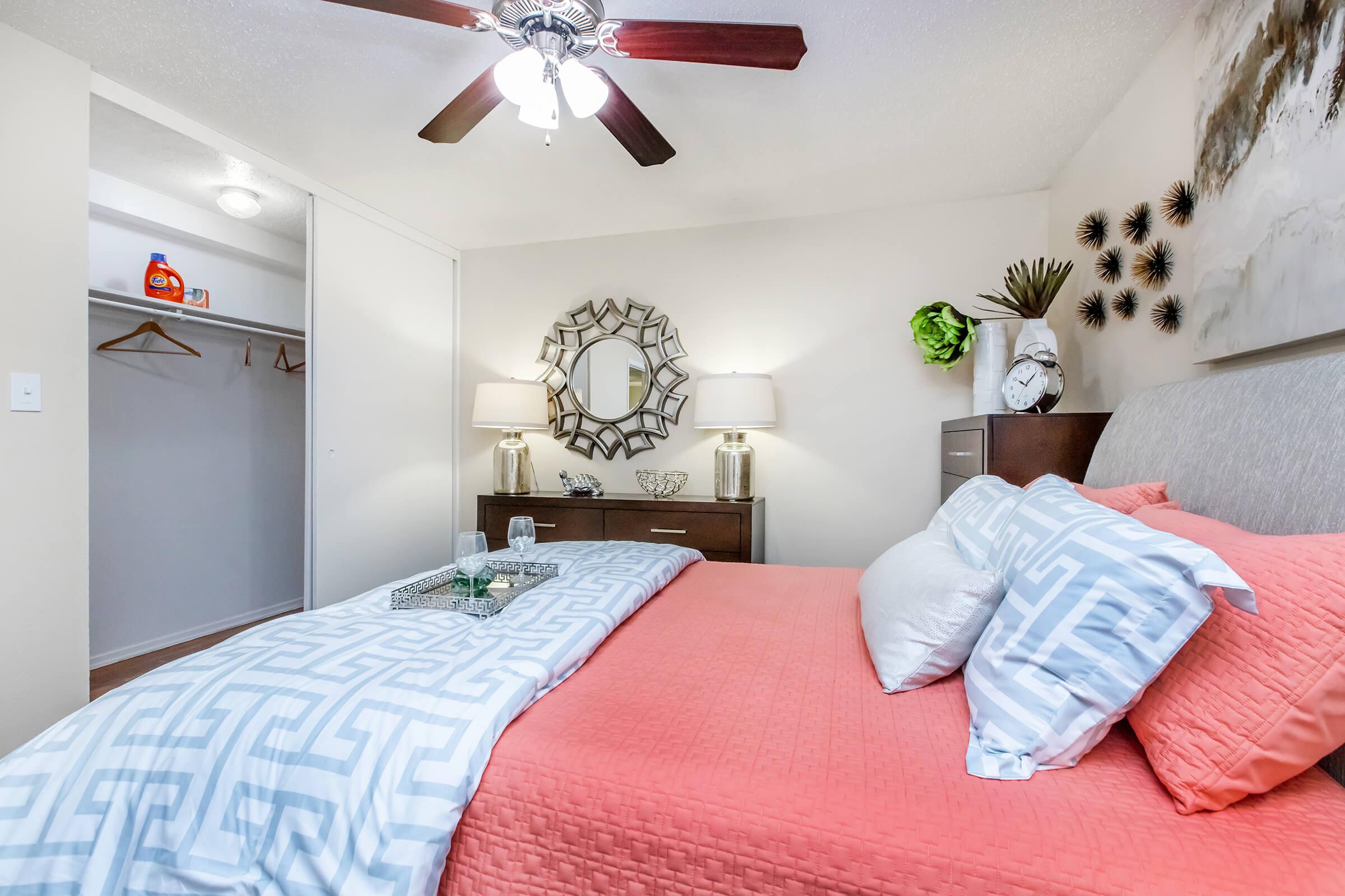 A cozy bedroom featuring a neatly made bed with coral and white bedding, a decorative mirror on the wall, two bedside lamps on a dark wood dresser, and a small plant. There is a closet with empty hangers visible in the background, creating a welcoming and inviting atmosphere.