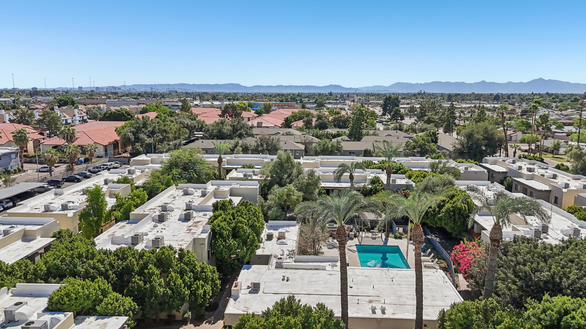 Aerial view of a suburban area featuring low-rise buildings and residential homes surrounded by trees. A swimming pool is visible in the center, with palm trees nearby. The background shows distant mountains under a clear blue sky.