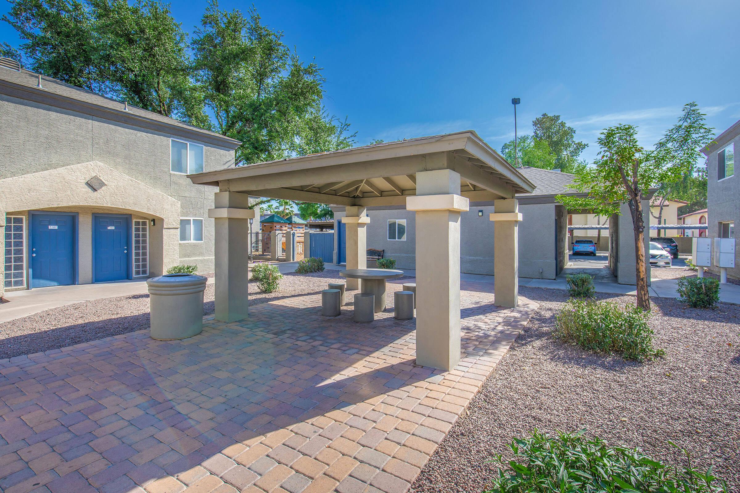 A shaded gazebo with a stone table and benches sits in a landscaped area surrounded by gravel and bushes. In the background, there are apartment buildings with blue doors and trees under a clear blue sky.