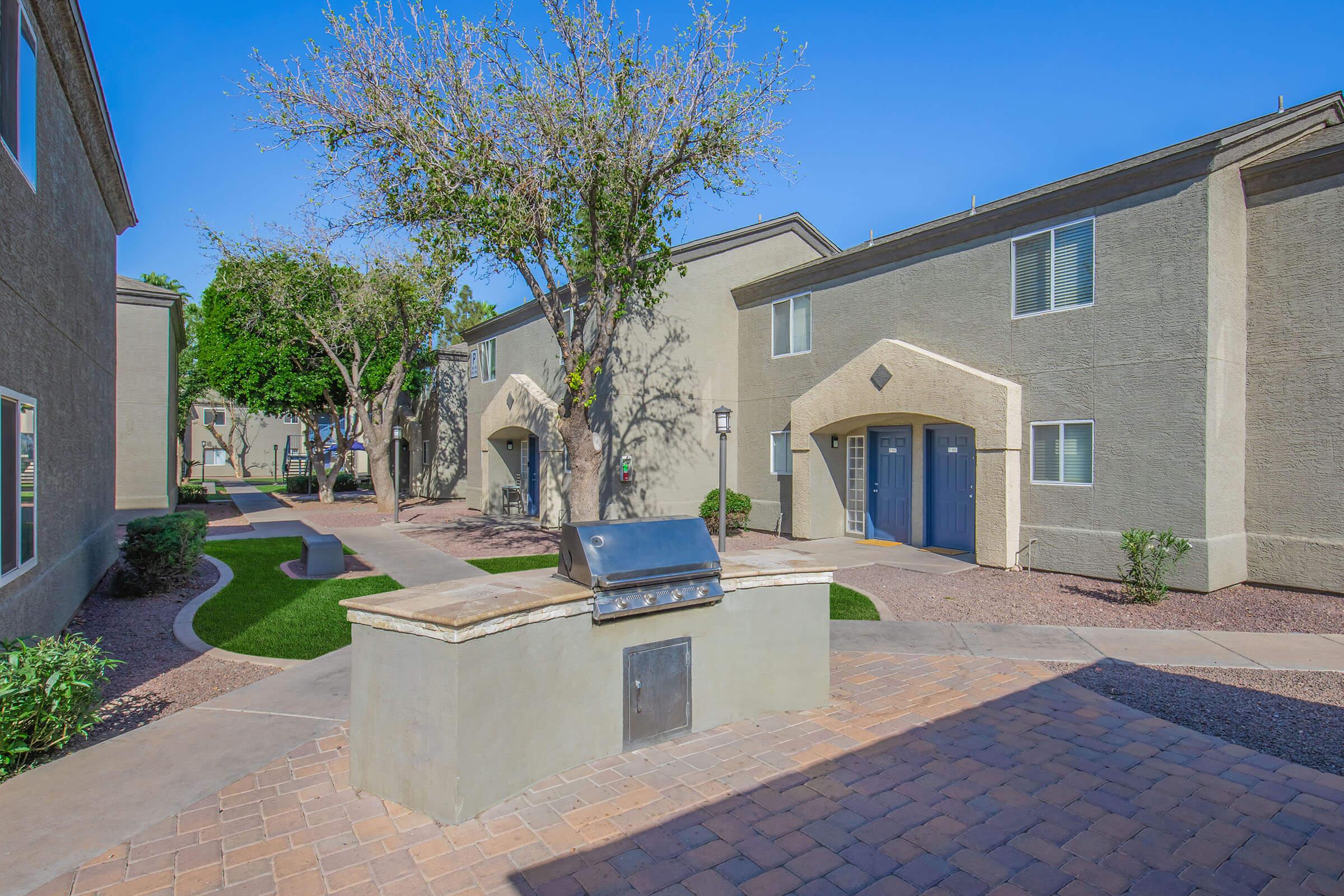 A view of a residential complex featuring two-story buildings with gray exteriors. In the foreground, there is a grill on a stone platform surrounded by landscaped paths and greenery. Trees are visible, and the sky is clear and blue, creating a peaceful outdoor setting.