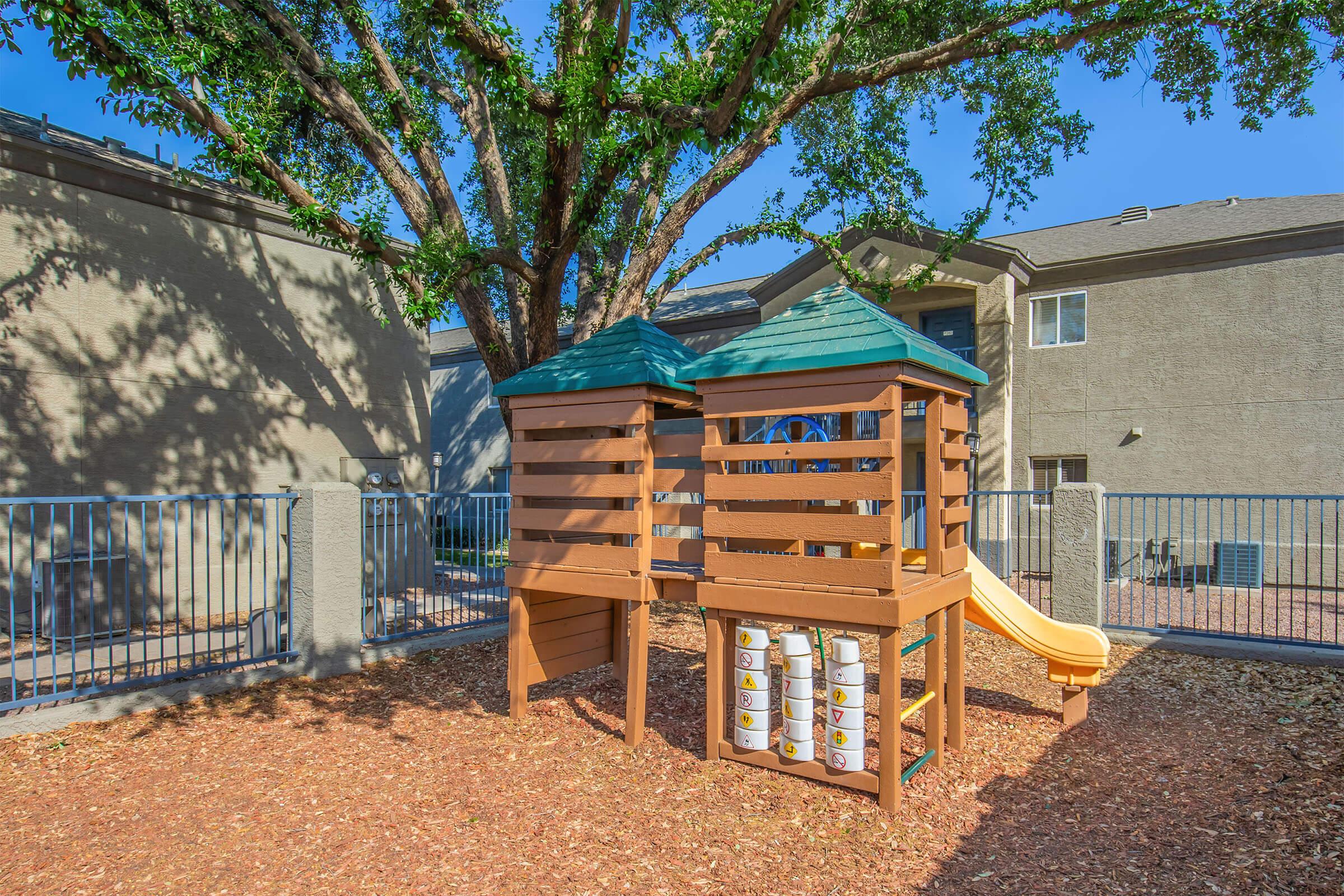 A wooden playground structure with a slide, situated in a fenced area with mulch beneath it. The structure features two towers with green roofs, surrounded by trees and a residential building in the background. The scene is under bright blue skies, creating a cheerful atmosphere for outdoor play.
