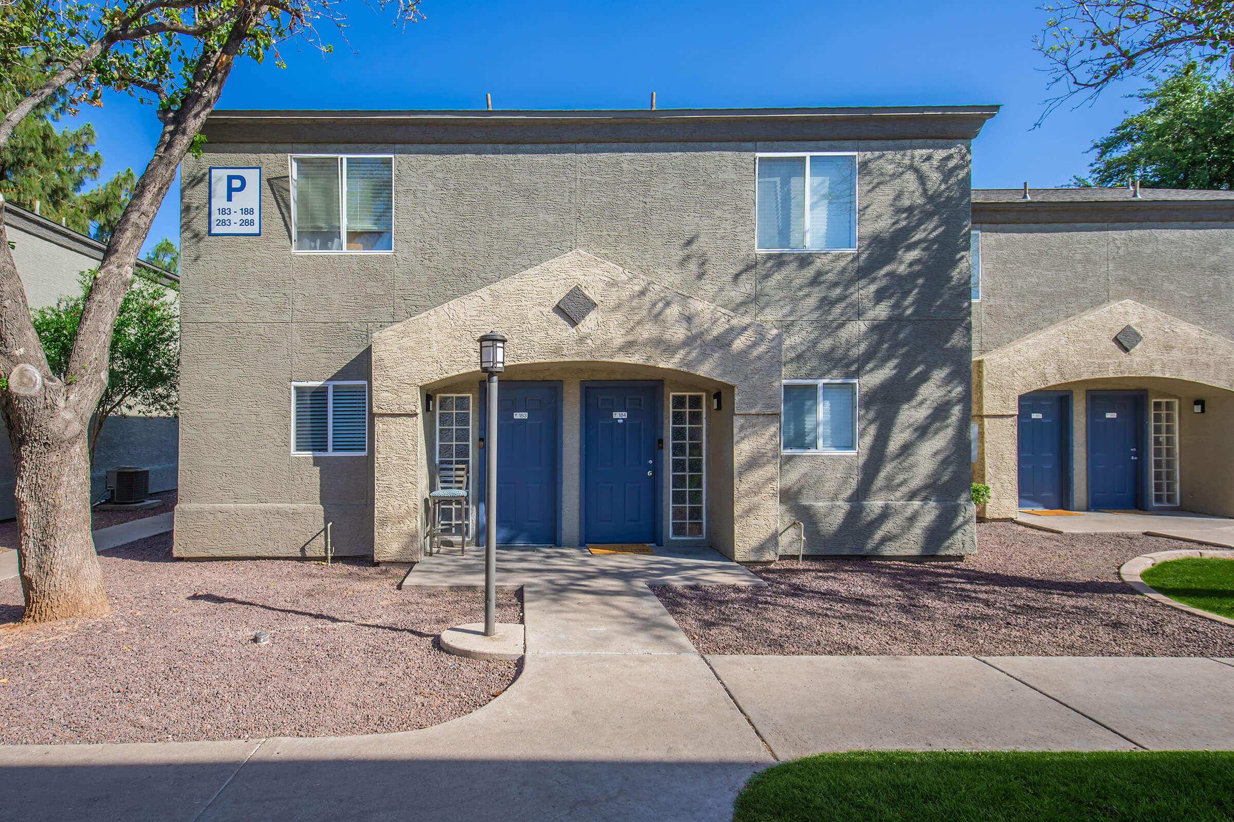 A gray two-story apartment building with a landscaped entrance. The unit doors are framed by arches, and there are parking signs visible above. The pathway is bordered by low-maintenance landscaping, and the setting is bright and sunny with clear blue skies.