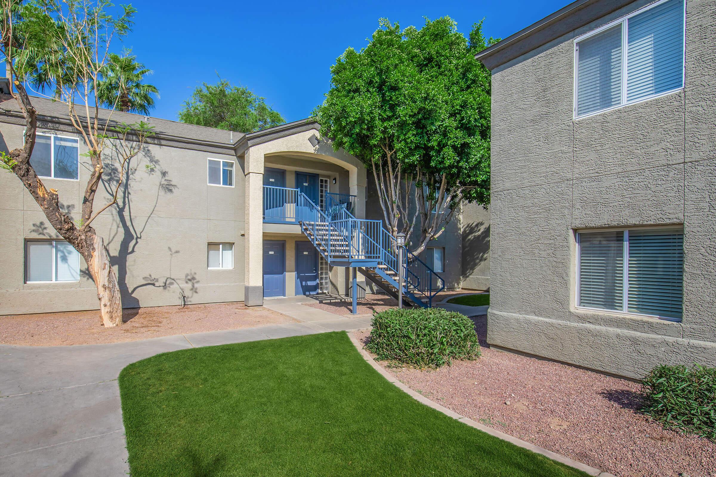 A view of an apartment complex featuring two buildings with light gray exteriors, a staircase leading to an upper level, and palm trees. The landscaping includes green grass and gravel, under a clear blue sky.
