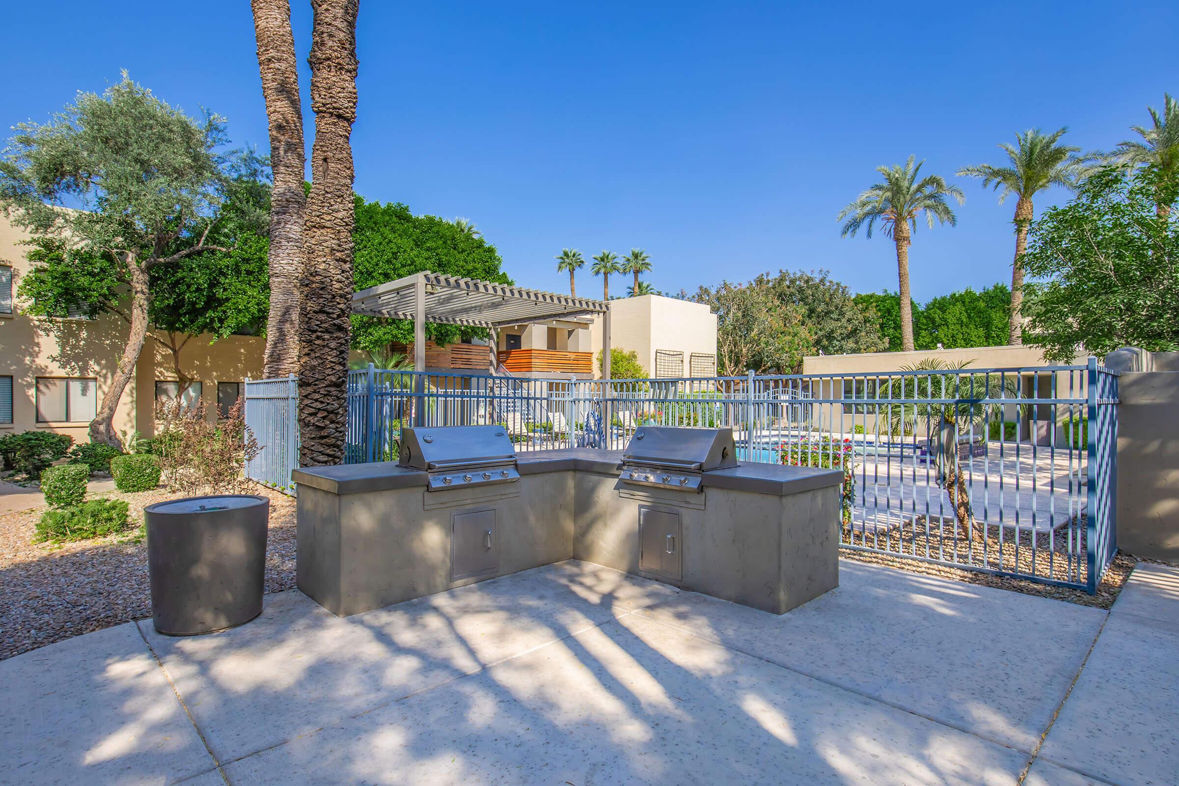 Outdoor grilling area featuring two built-in barbecue grills, surrounded by palm trees and landscaped greenery. A gated pool area is visible in the background, under a clear blue sky. The setting is designed for relaxation and social gatherings.