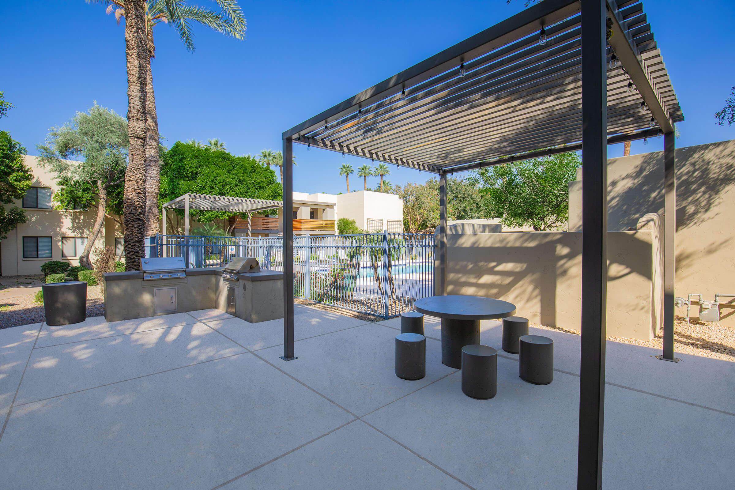 A modern outdoor patio area featuring a shaded pergola with a circular table and seating. Nearby, there is a barbecue grill, surrounded by a landscaped garden with palm trees and greenery. In the background, a swimming pool is visible, creating a relaxing atmosphere for gatherings and leisure.