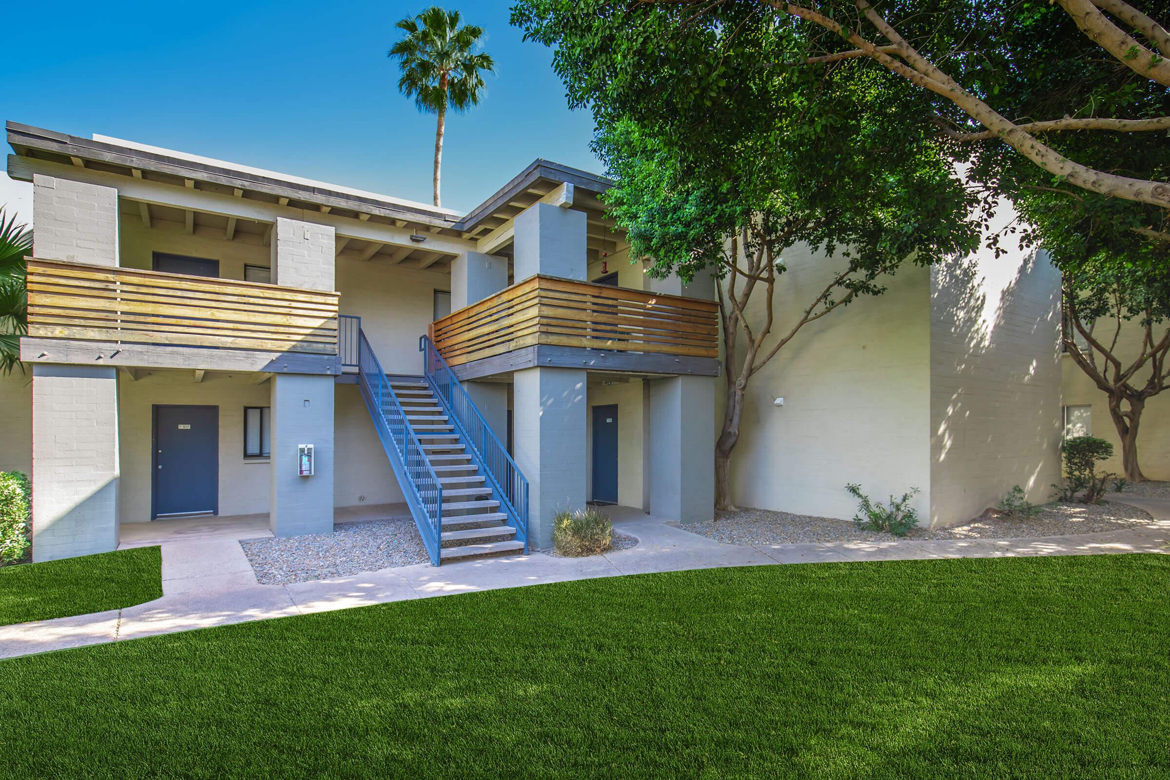 Two-story building with a modern design featuring a wooden balcony and stairs. It is surrounded by lush green grass, palm trees, and landscaped shrubs. The exterior walls are painted in light tones, and there are doorways with gray doors leading to the apartments. The scene is set under a clear blue sky.