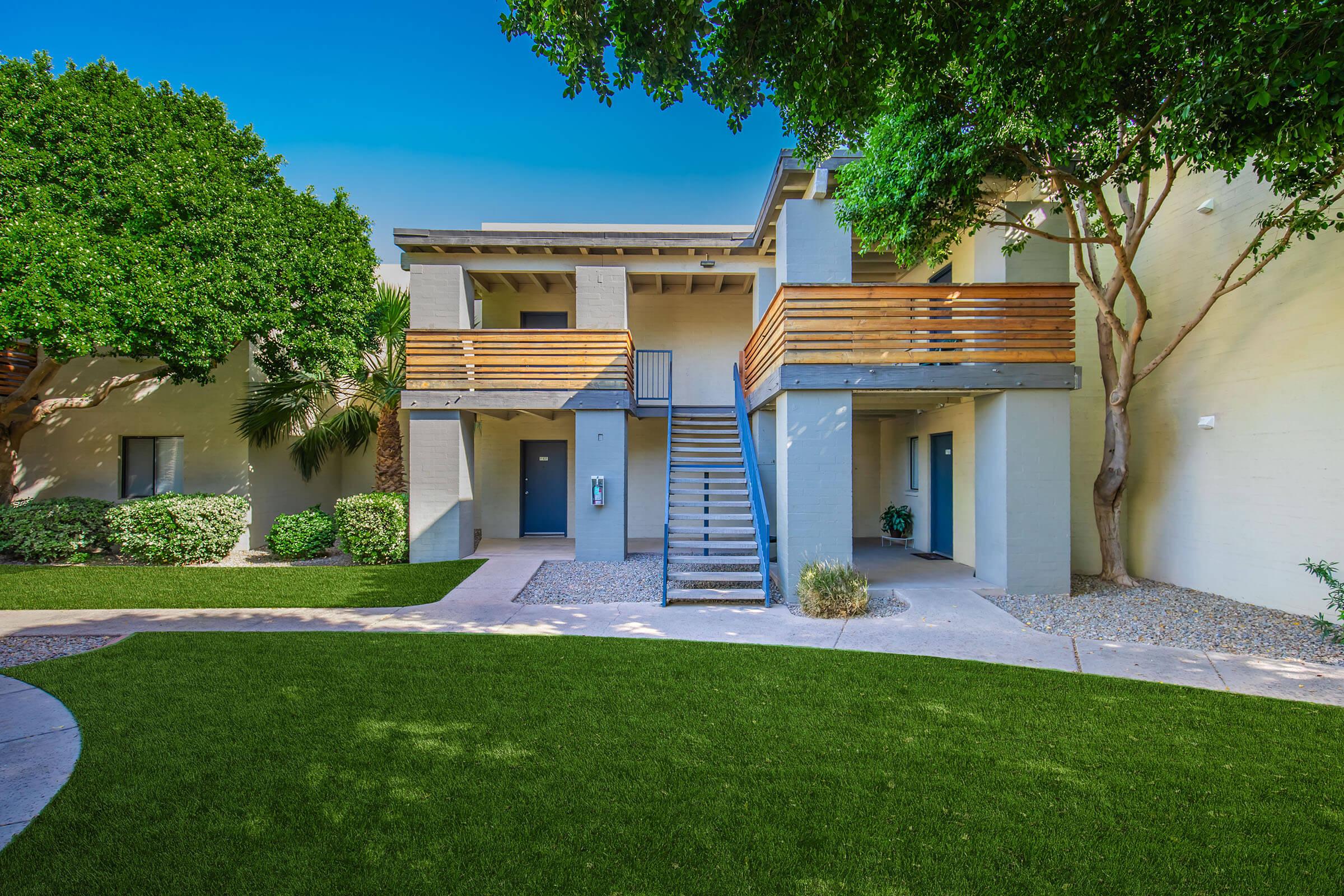 Two-story building surrounded by green landscaping, featuring a staircase leading to a balcony on the second floor. The building has a light-colored exterior with wooden railing and is set against a backdrop of trees. The area includes well-maintained grass and pathways.