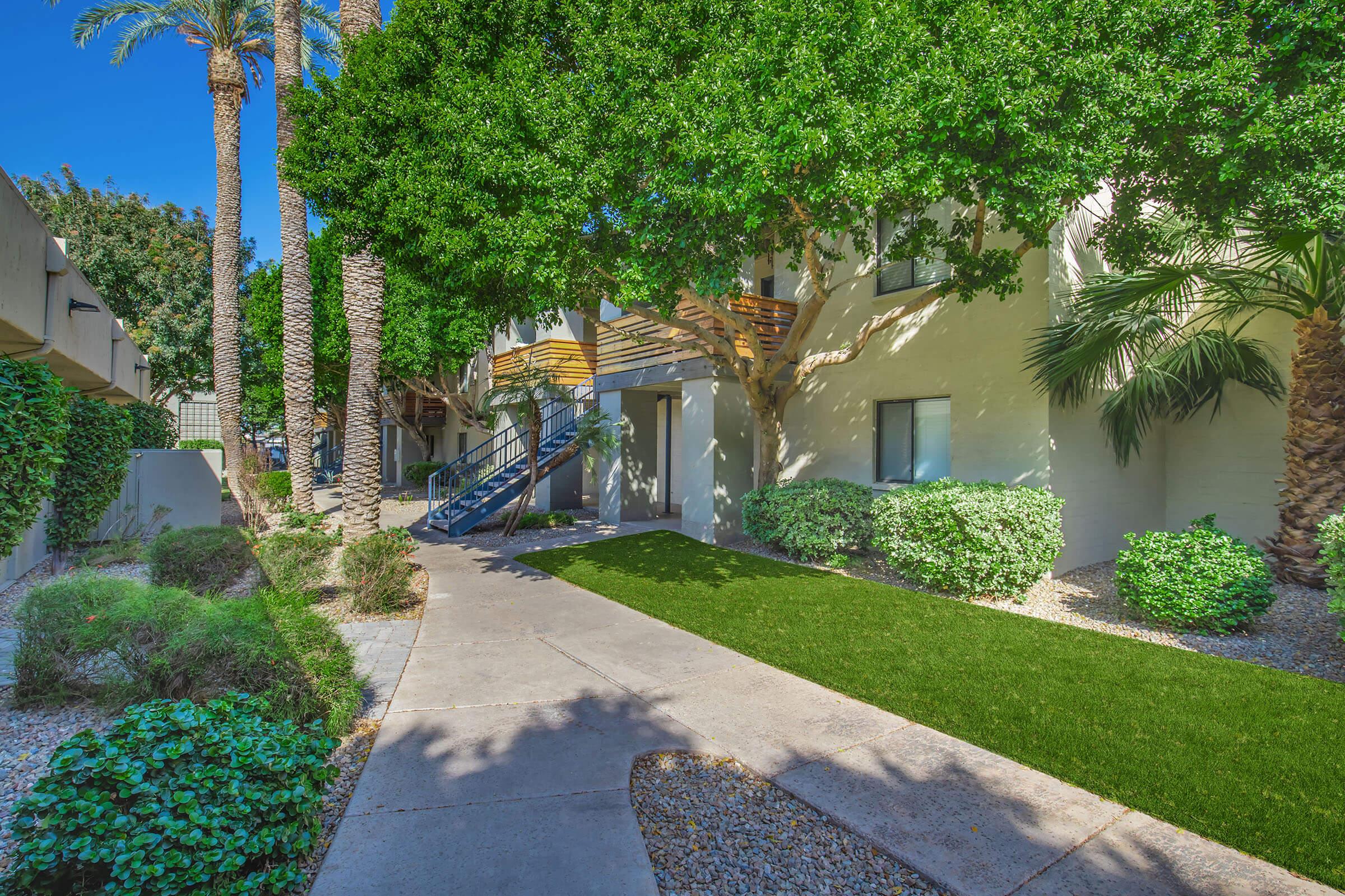 A well-maintained pathway leading to a two-story apartment building, surrounded by lush greenery, palm trees, and decorative bushes. The area features a small patch of artificial grass and a staircase leading to the upper floor, creating a warm and inviting atmosphere. Sunlight casts gentle shadows on the ground.