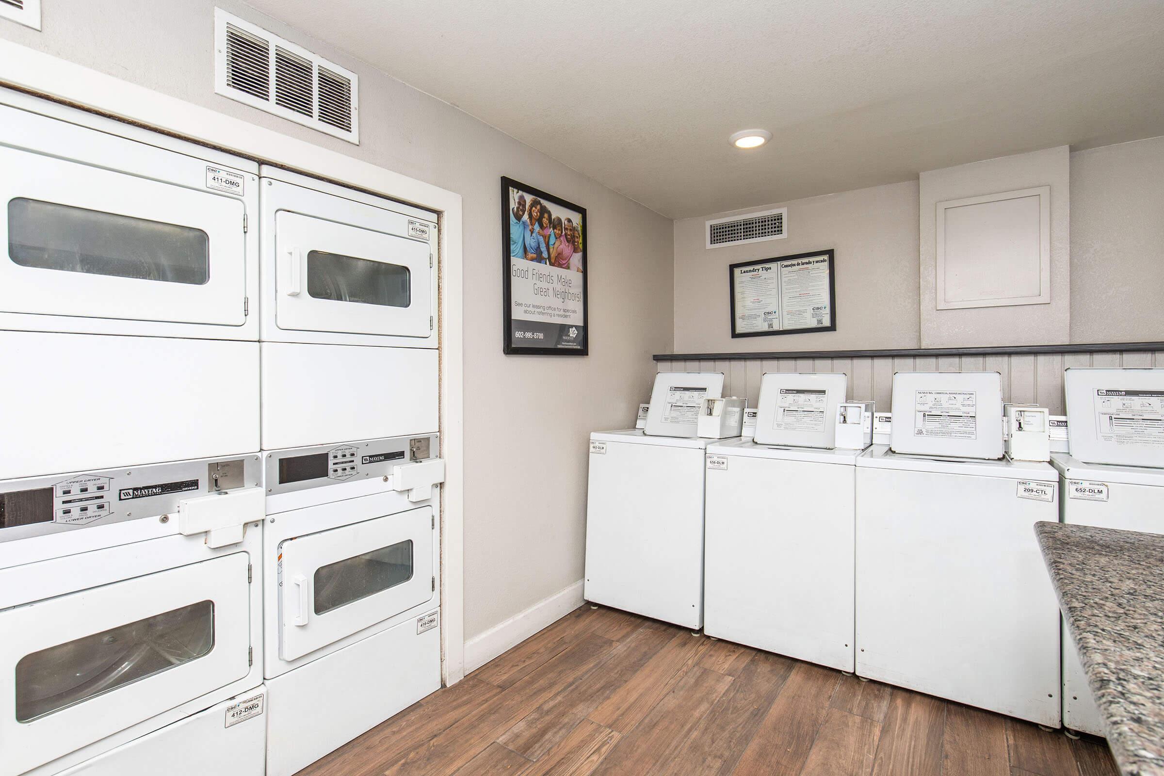 A laundry room featuring stacked washing machines and dryers on the left, and standalone washers and dryers on the right. The room has light-colored walls, wooden flooring, and a bulletin board with posters or information on the wall. Bright lighting enhances the overall space.