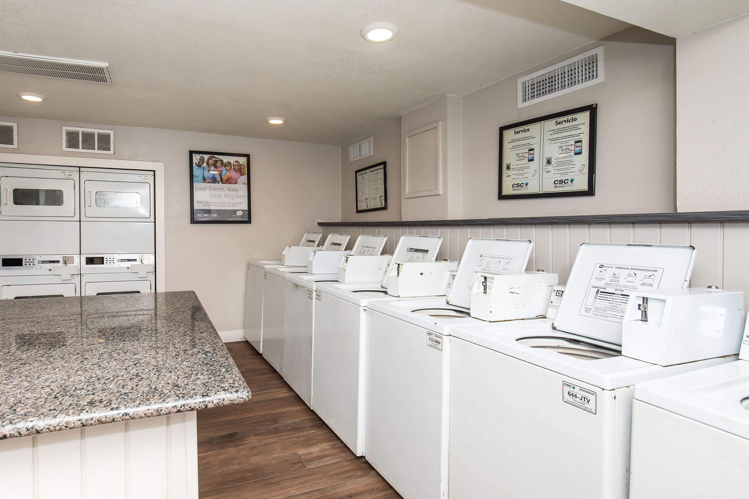 A clean and modern laundry room featuring several white washing machines and dryers against a wall. There is a granite countertop in the foreground, and informational posters and service listings are displayed on the walls. The flooring is wooden, and the space is well-lit and organized.