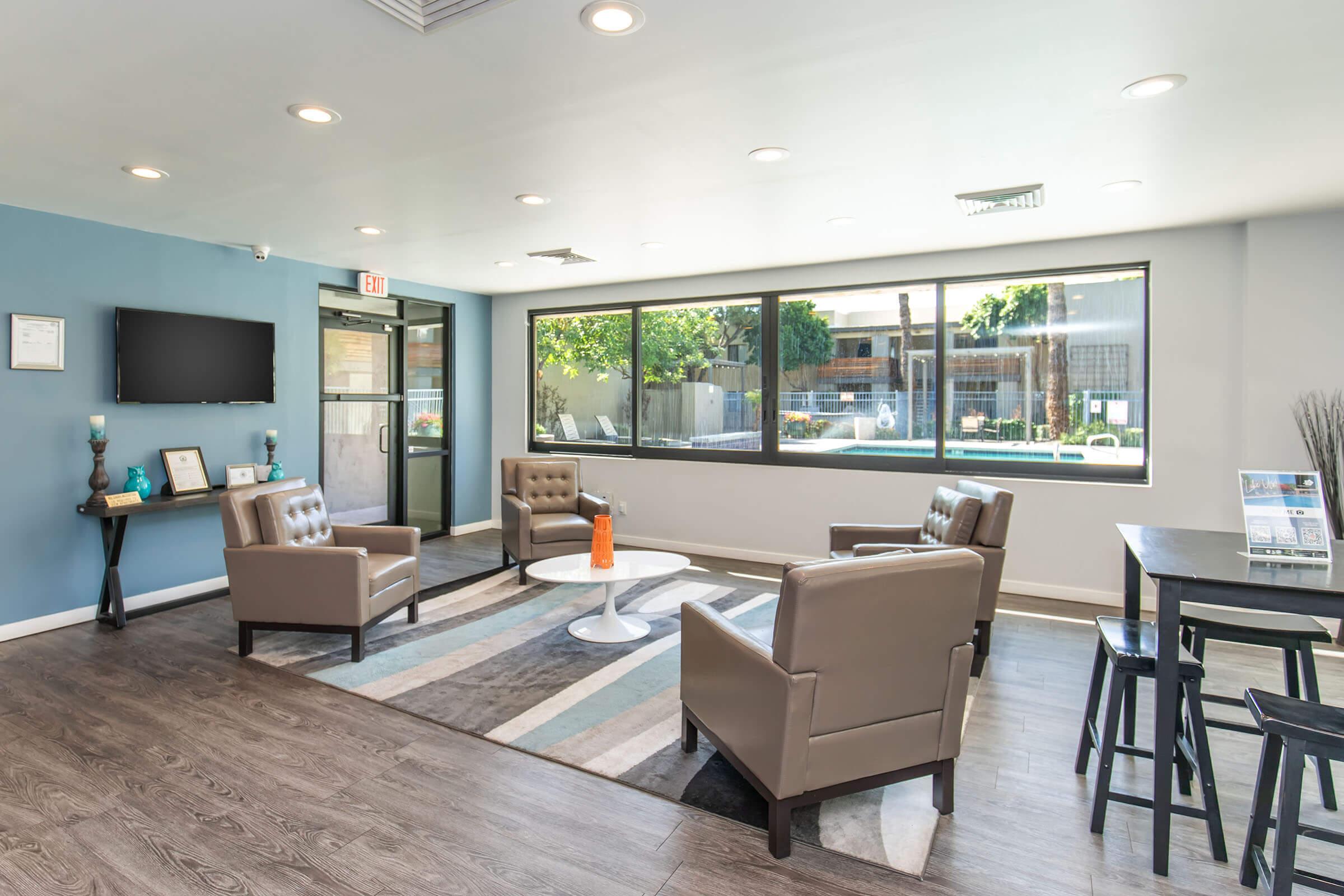 A modern lounge area featuring two brown leather armchairs, a white coffee table, and a rug with blue and gray stripes. A flat-screen TV is mounted on the wall, and there's a dining table with chairs. Large windows let in natural light, showing green outdoor landscaping.