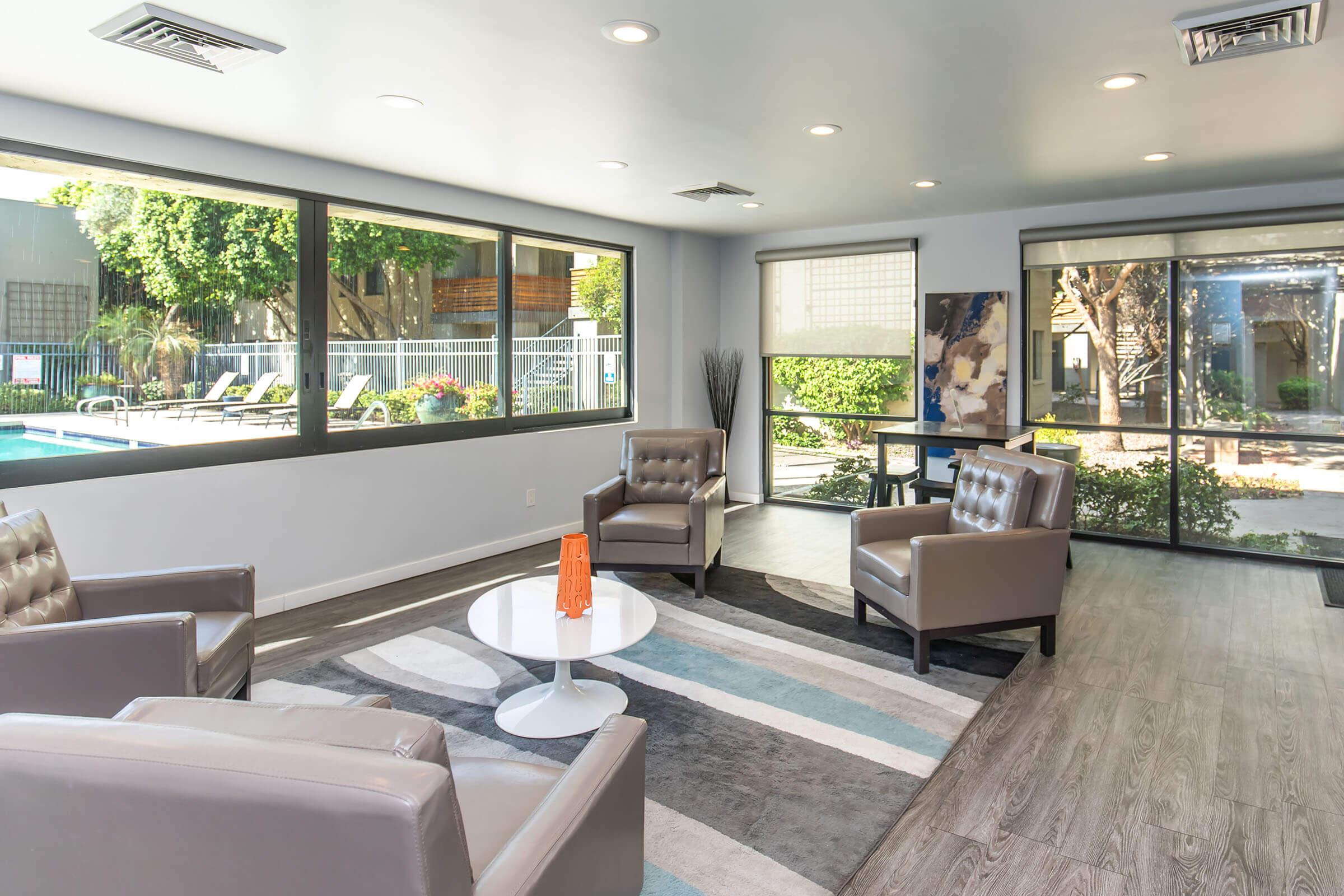 Bright and modern indoor lounge area featuring two gray armchairs, a round white coffee table, and a colorful rug. Large windows offer views of a landscaped outdoor area with a swimming pool. The space is well-lit, with potted plants adding greenery, creating a welcoming atmosphere.
