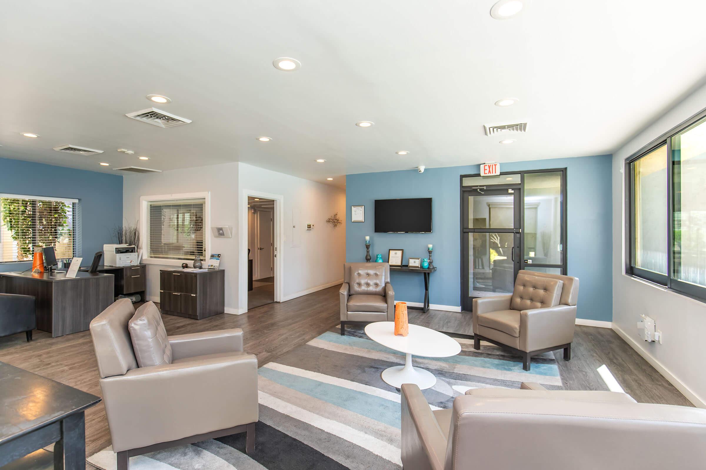 Modern waiting area featuring cozy taupe armchairs and a round white coffee table on a striped rug. The walls are painted in calming shades of blue and light gray. There's a reception desk visible in the background, along with a large window allowing natural light in, creating an inviting atmosphere.