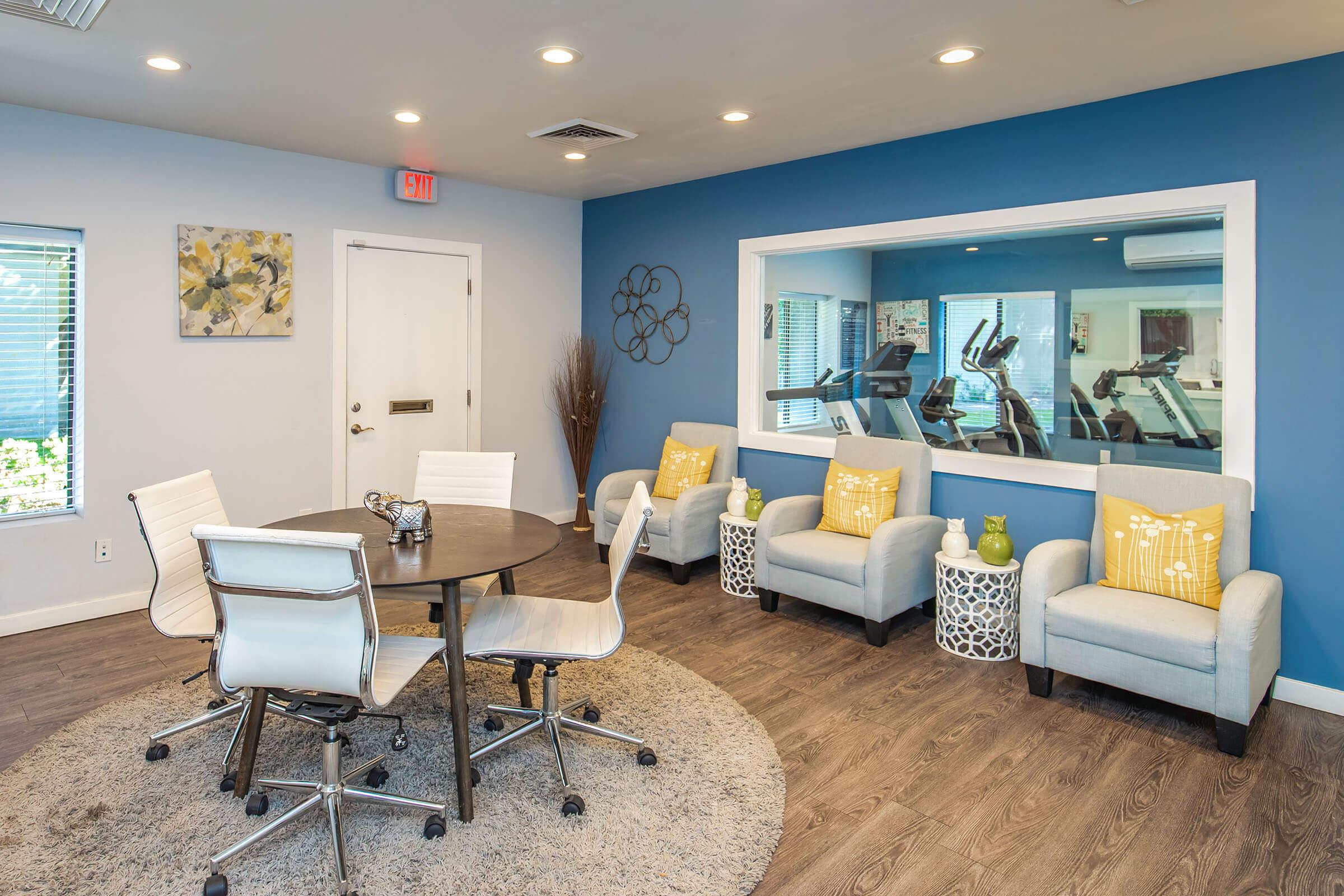 A modern lounge area featuring a circular table with four rolling chairs, two light blue armchairs with yellow cushions, and a round rug on wooden flooring. A large window looks into a fitness area, and complemented by decorative wall art and plants. The walls are painted in a calming blue tone.