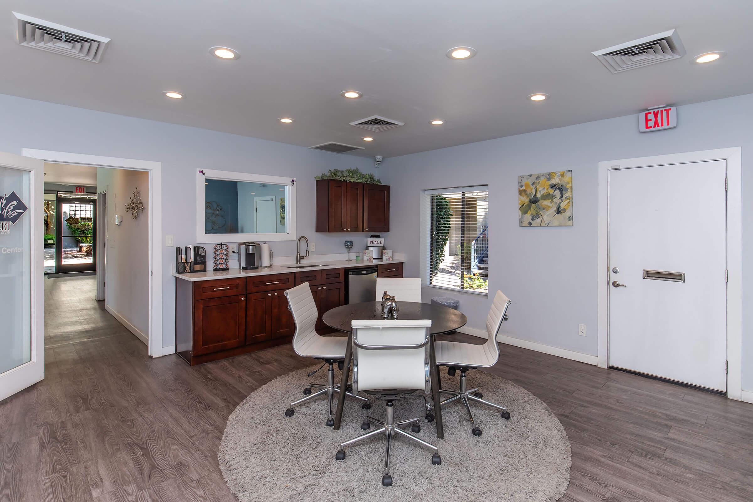 A modern kitchen and dining area featuring a round table with four chairs, dark wood cabinets, and stainless steel appliances. The walls are painted light blue, and there are large windows allowing natural light. A rug under the table adds warmth to the space, with an exit door visible on the right.
