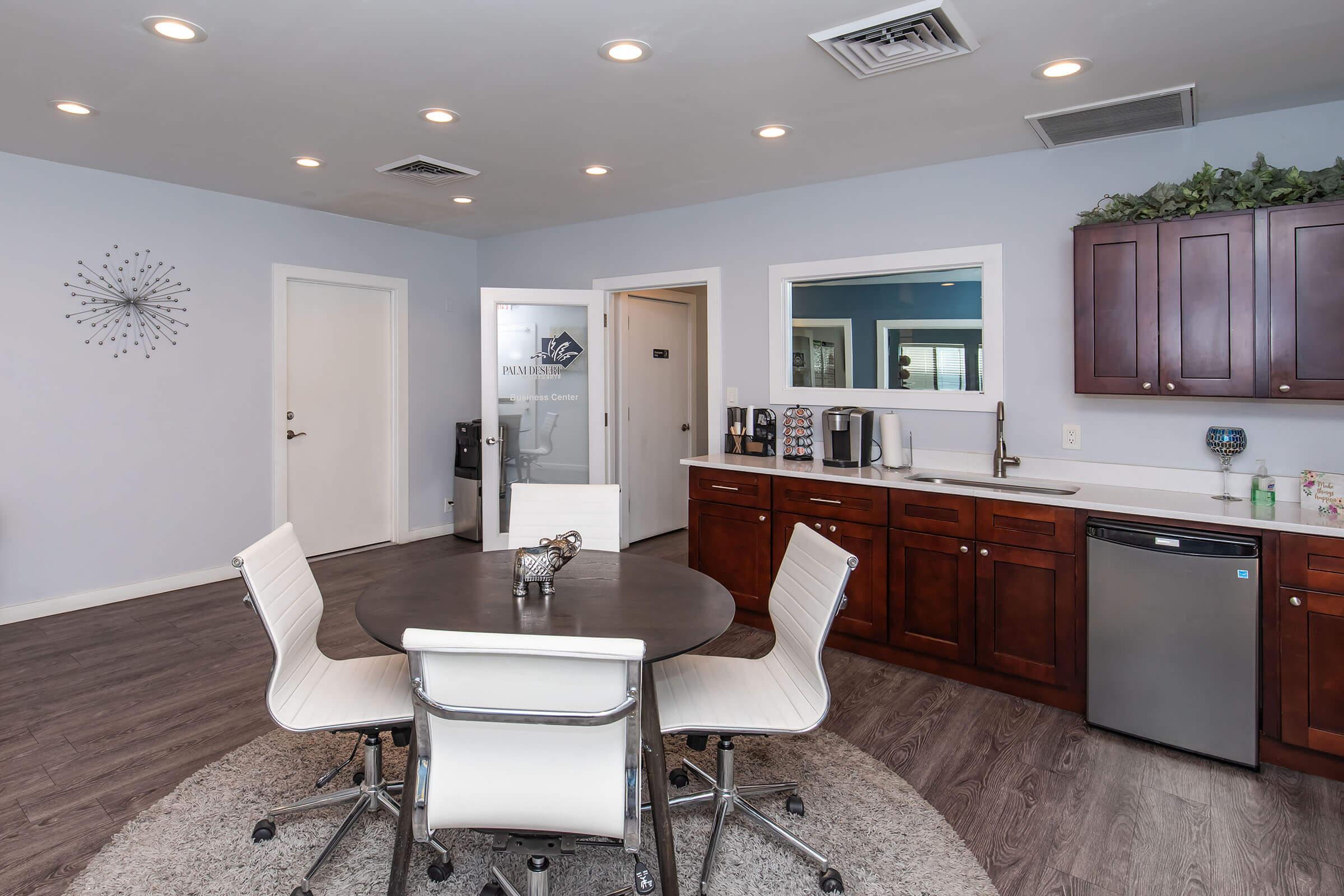 A modern kitchenette featuring a round dining table with white chairs, dark wood cabinetry, a small refrigerator, a coffee machine, and a decorative rug. The walls are painted light blue, and there are doors leading to other rooms. Ceiling lights provide bright illumination, enhancing the inviting atmosphere.