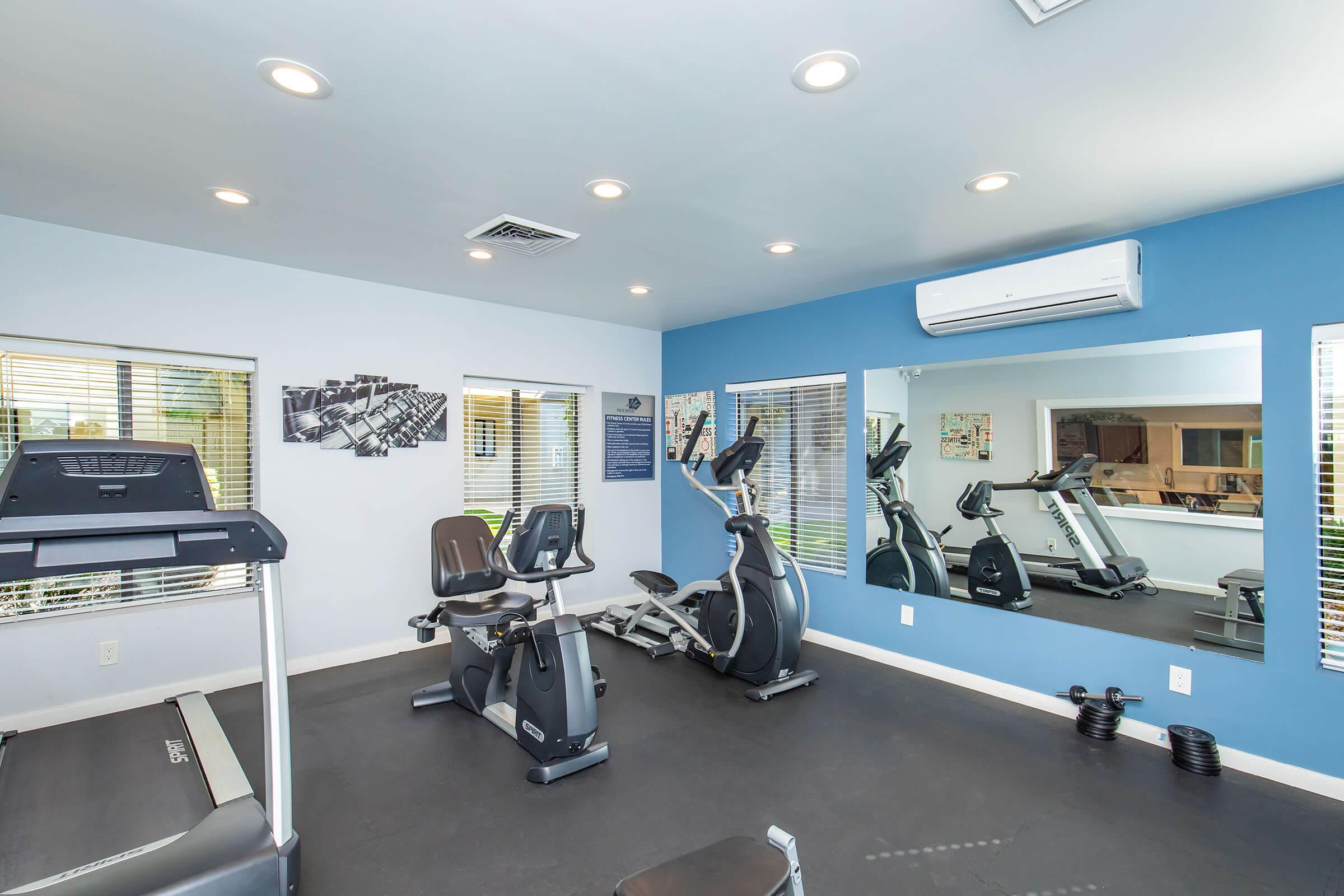 A modern gym interior featuring exercise equipment, including a treadmill and stationary bike. The walls are painted blue, and there are large windows with blinds allowing natural light. Mirrors are positioned to reflect the equipment, creating an open and spacious feel.