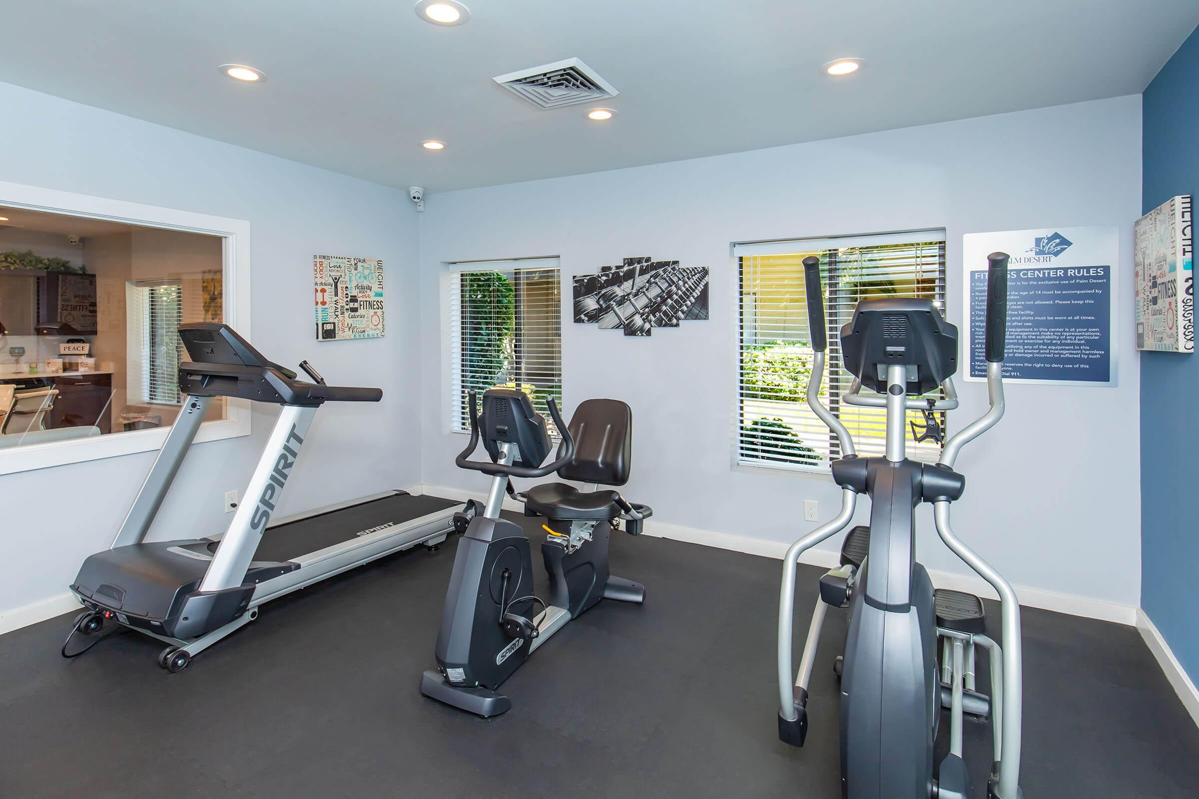 A well-lit fitness room featuring a treadmill, an exercise bike, and an elliptical machine. The walls are painted light blue, with a graphic display of exercise rules and a window offering natural light. The space is designed for workouts and physical fitness activities.