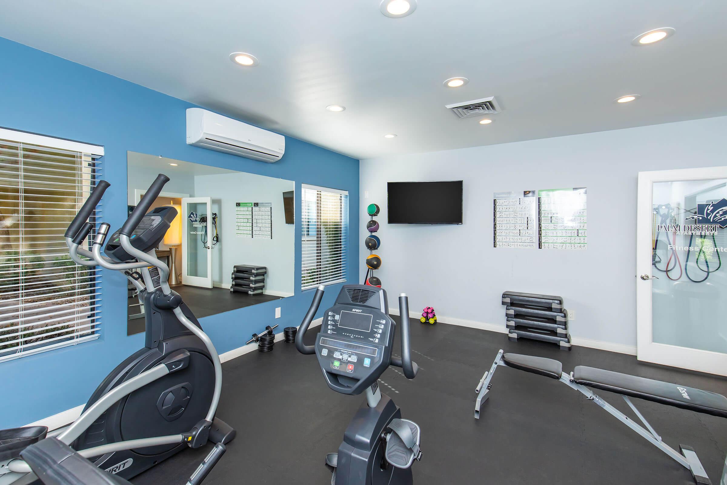 A modern gym interior featuring an elliptical machine, a weight bench, and free weights on racks. There are mirrors on the wall, a flat-screen TV, and bright lighting. The room is painted blue with white accents, creating a clean and inviting workout space.