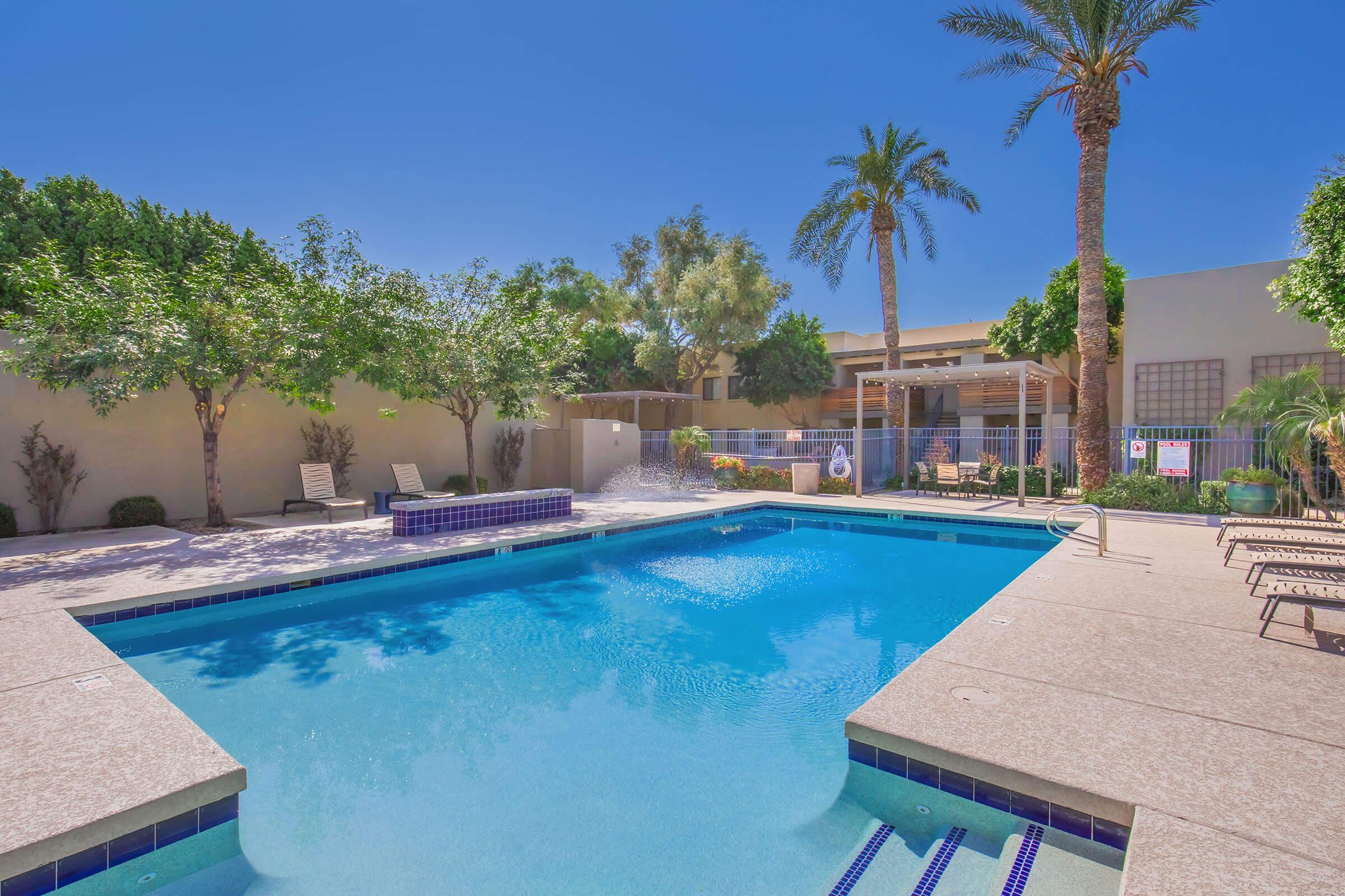 A clear blue swimming pool surrounded by palm trees and greenery. Lounge chairs are positioned around the pool area, and a small fountain is visible. The sky is bright and sunny, creating a cheerful and inviting atmosphere.