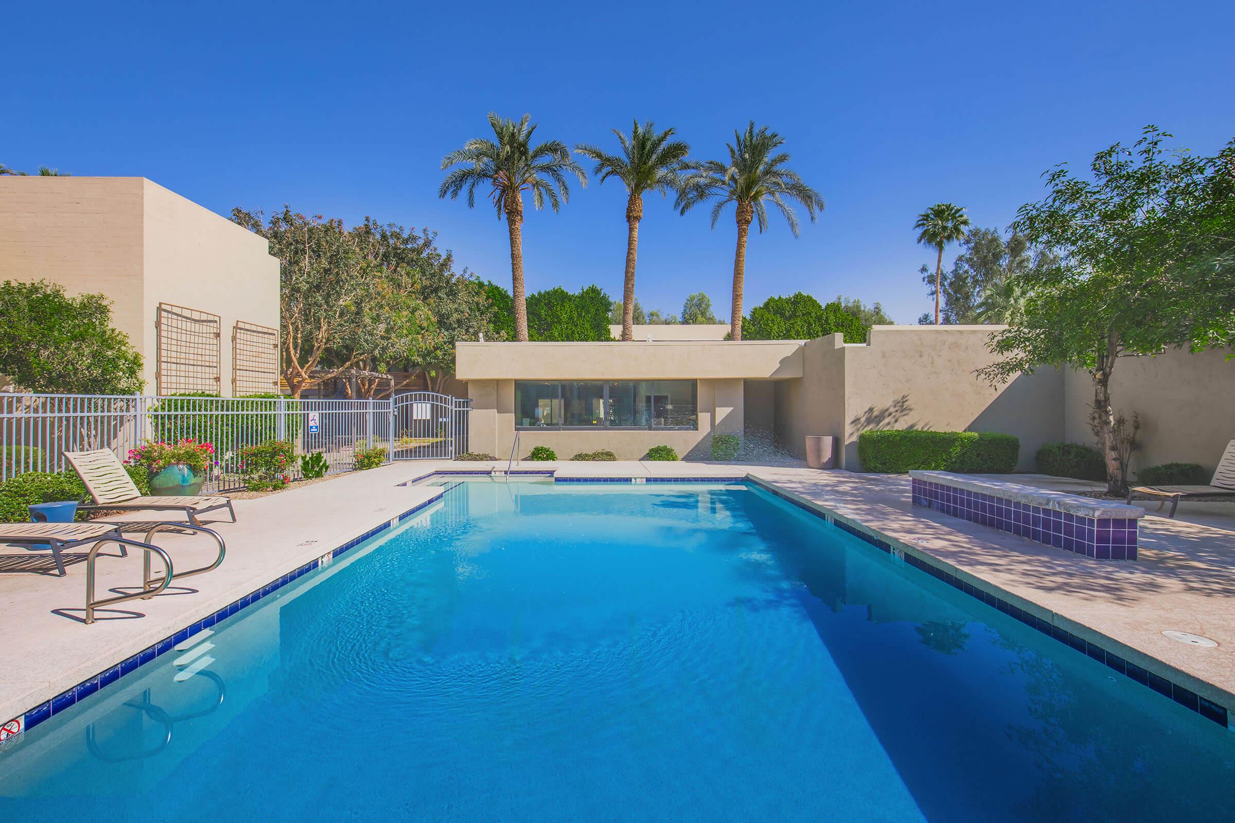 A serene outdoor pool surrounded by palm trees and landscaping. The pool features a clear blue water surface with a small spa area to one side. Modern architecture is visible in the background, along with lounge chairs and a fence enclosing the area. The scene is bright and inviting, set against a clear blue sky.