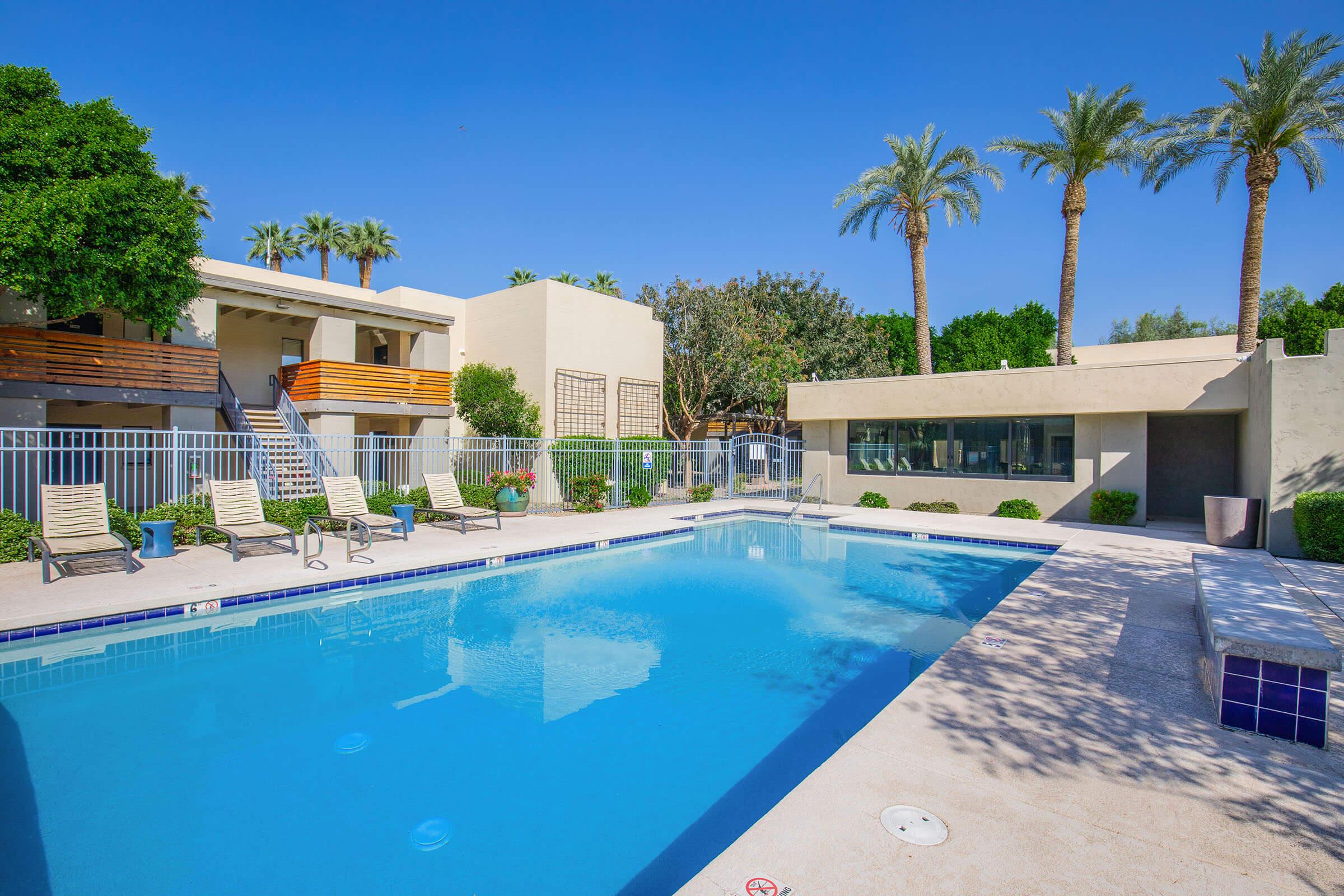 A clear blue swimming pool surrounded by palm trees and lounge chairs. In the background, a modern building with greenery and a spacious area. Bright and sunny atmosphere, perfect for relaxation.